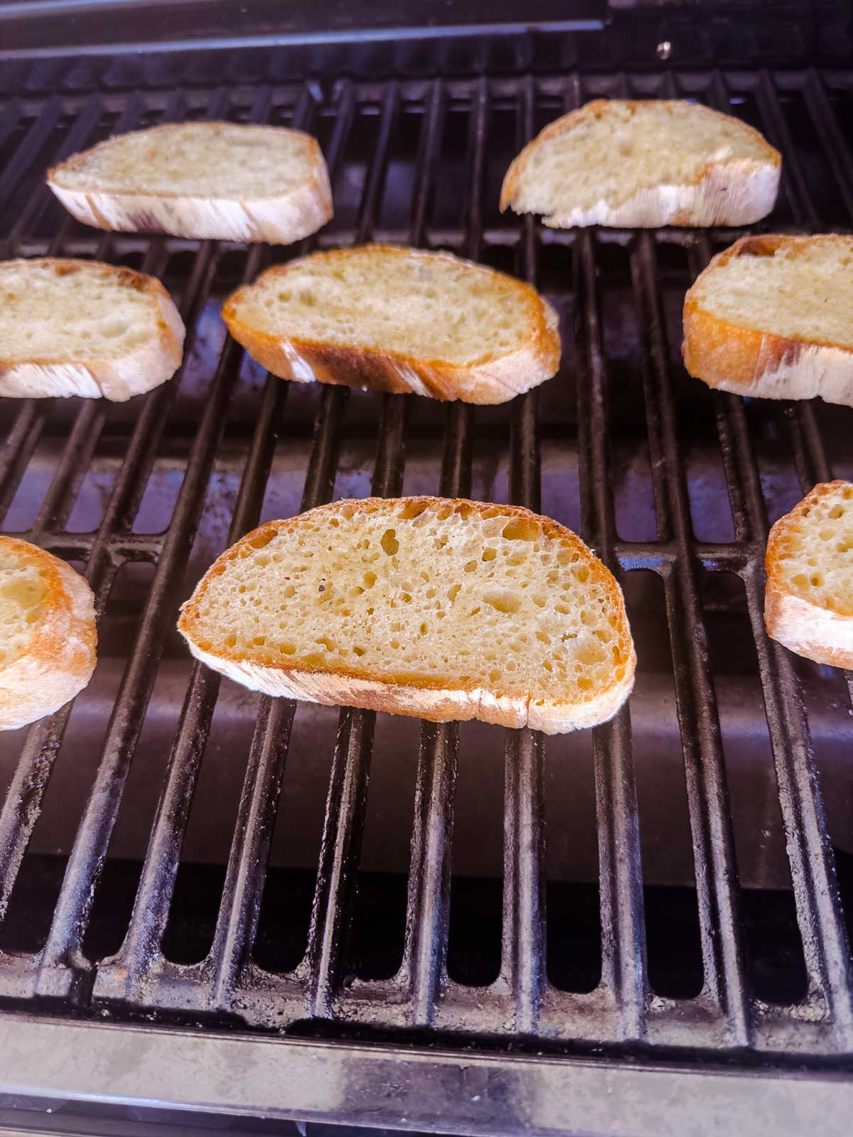 Several slices of rustic bread are toasting on a grill, with grill marks beginning to form on the undersides. The bread slices are evenly spaced across the grates.