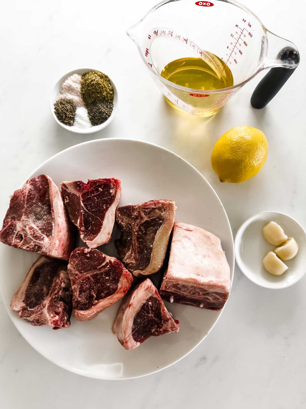 A plate of raw lamb loin chops, a small bowl with garlic cloves, a halved lemon, a dish of mixed spices, and a measuring cup of oil are arranged on a white surface.