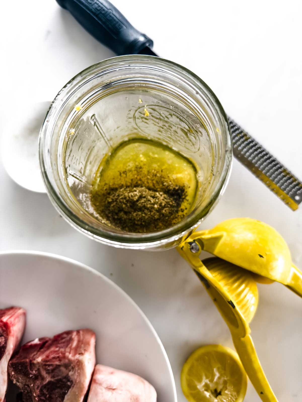 A glass jar with marinade, topped with spices, sits on a white surface near a lemon juicer with squeezed lemons, a zester, a spoon, and a plate with raw meat pieces.