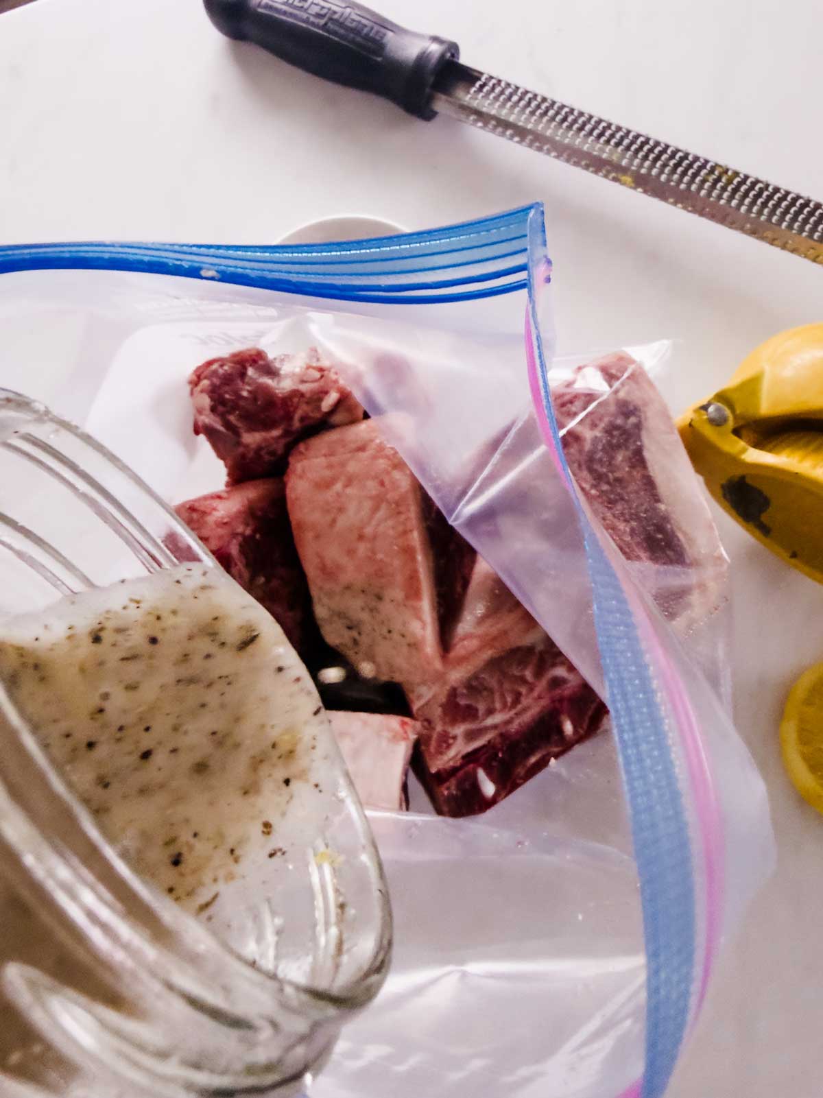Chunks of raw beef in a ziplock bag being marinated with a creamy sauce. A grater, a squeezed lemon half, and a whole lemon are visible on a white surface nearby.