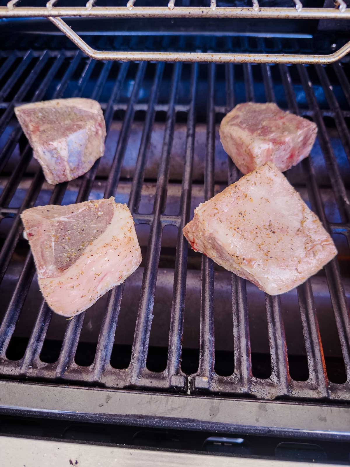 Four thick, seasoned pieces of raw beef are placed on a grill, ready to be cooked over the open flame. The metal grill grates and an upper rack are visible.