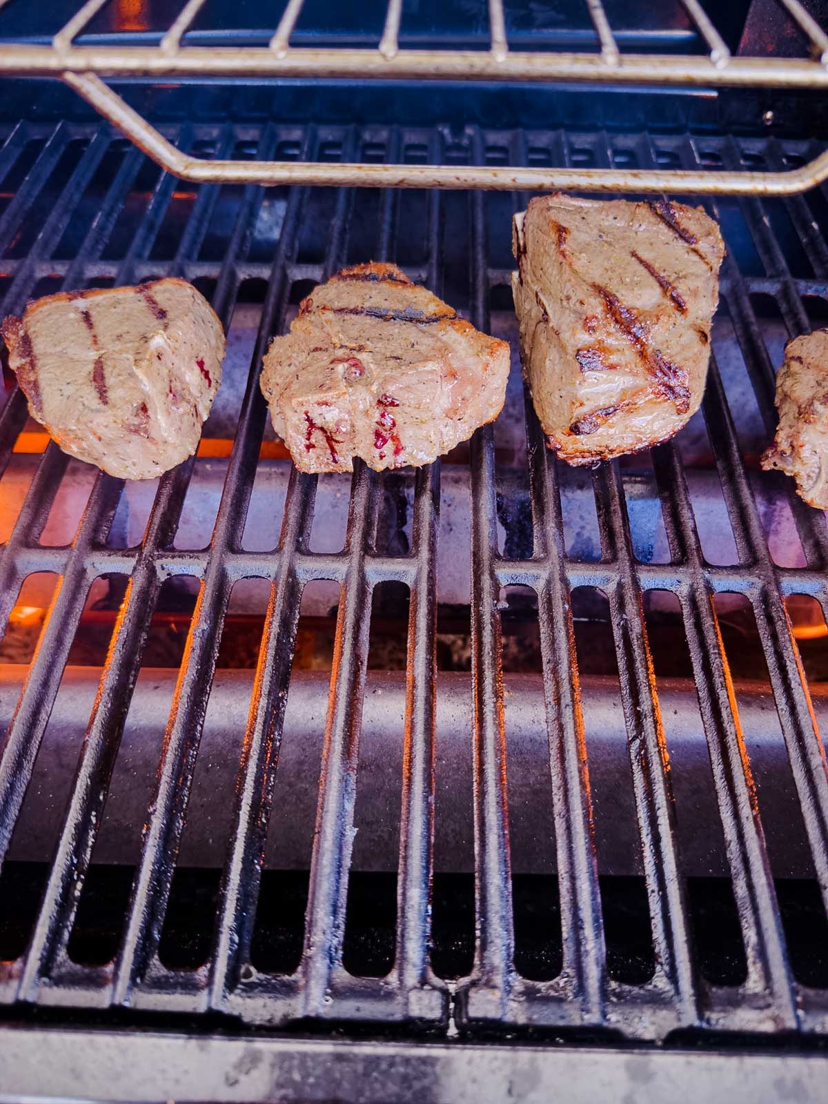Four pieces of steak with grill marks cooking on a barbecue grill over open flames.