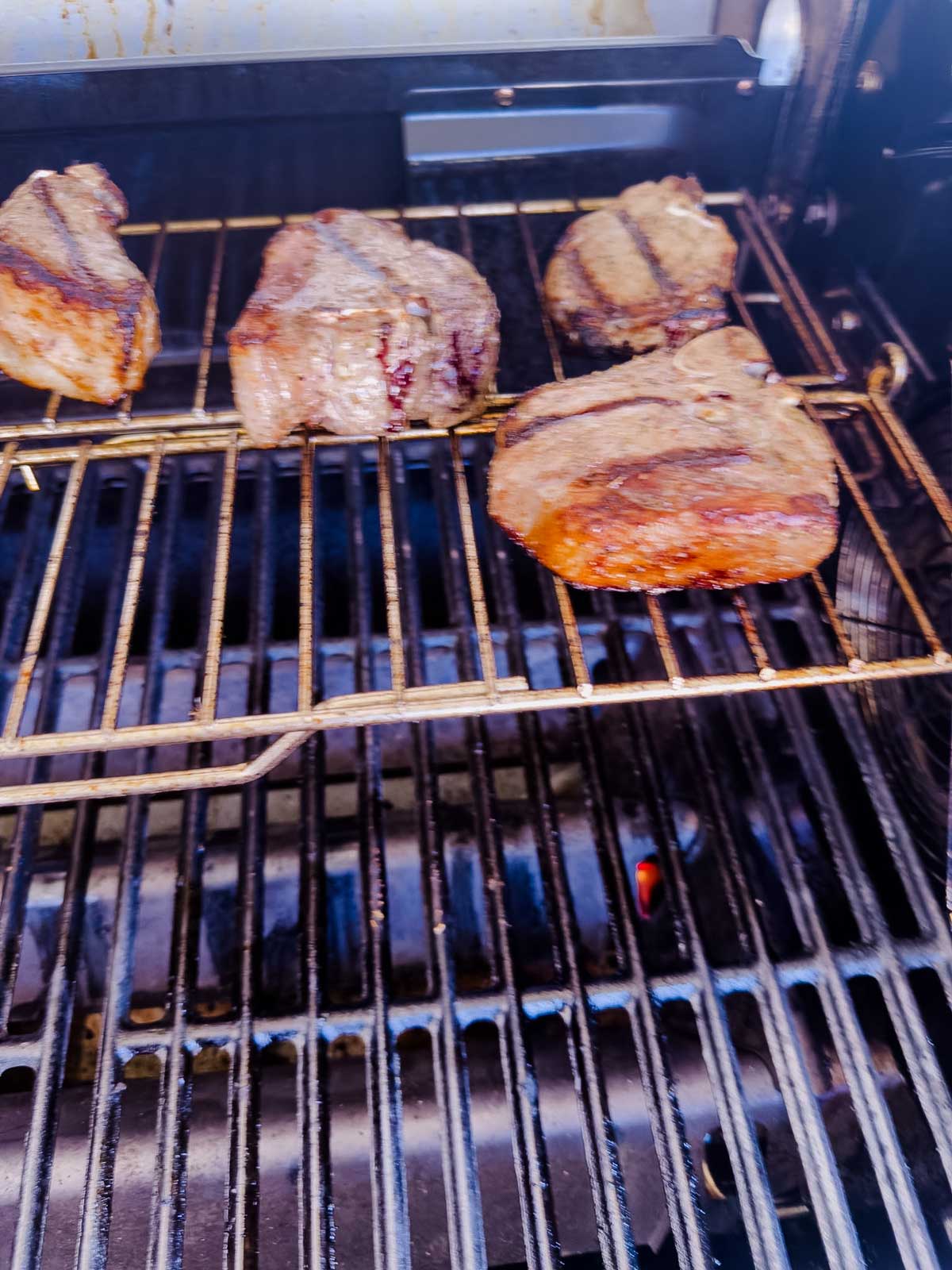 Four pieces of grilled steak are cooking on the upper rack of a barbecue grill, with grill marks visible on the meat and flames below the lower rack.