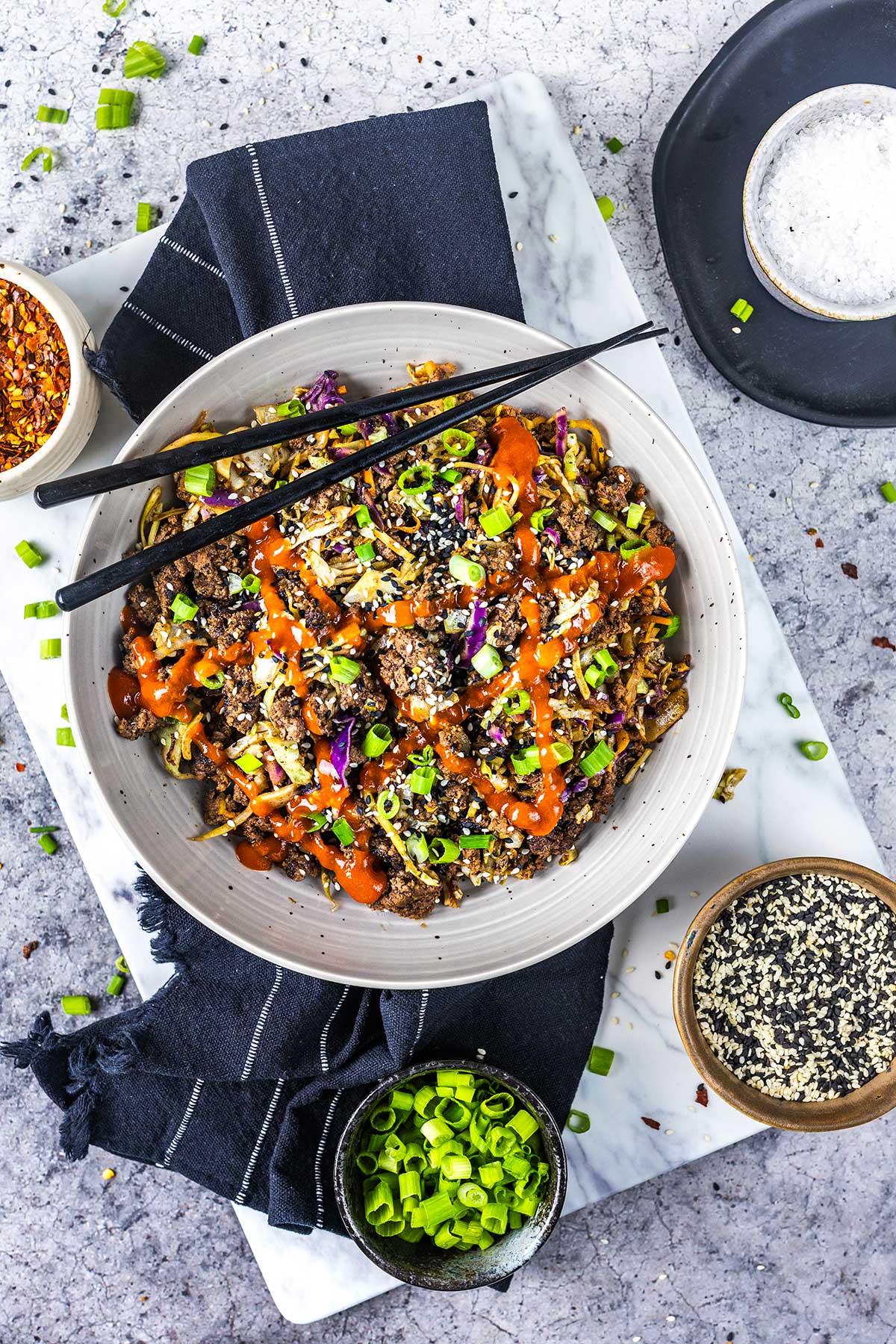 A bowl of noodles topped with ground meat, sliced green onions, sesame seeds, shredded vegetables, and a drizzle of bright orange sauce. Black chopsticks rest on the bowl, surrounded by small bowls of seasonings and garnishes.