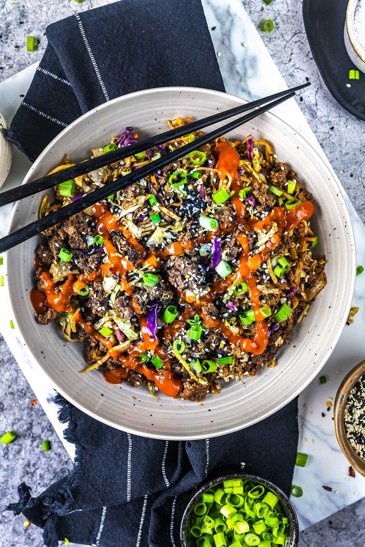 Egg roll in a bowl with ground beef, sliced green onions, sesame seeds, shredded cabbage, and a drizzle of bright orange sauce, with black chopsticks resting on the bowl.
