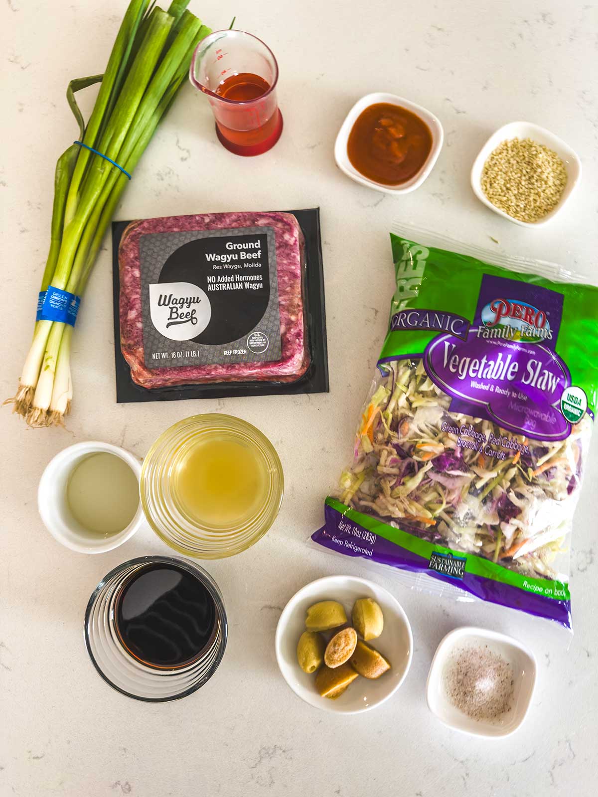 Ingredients for an Asian-inspired Blackstone egg roll in a bowl on a white surface: green onions, ground Wagyu beef, vegetable slaw mix, bowls with sauces and juice, green olives, sesame seeds, salt, and sugar.