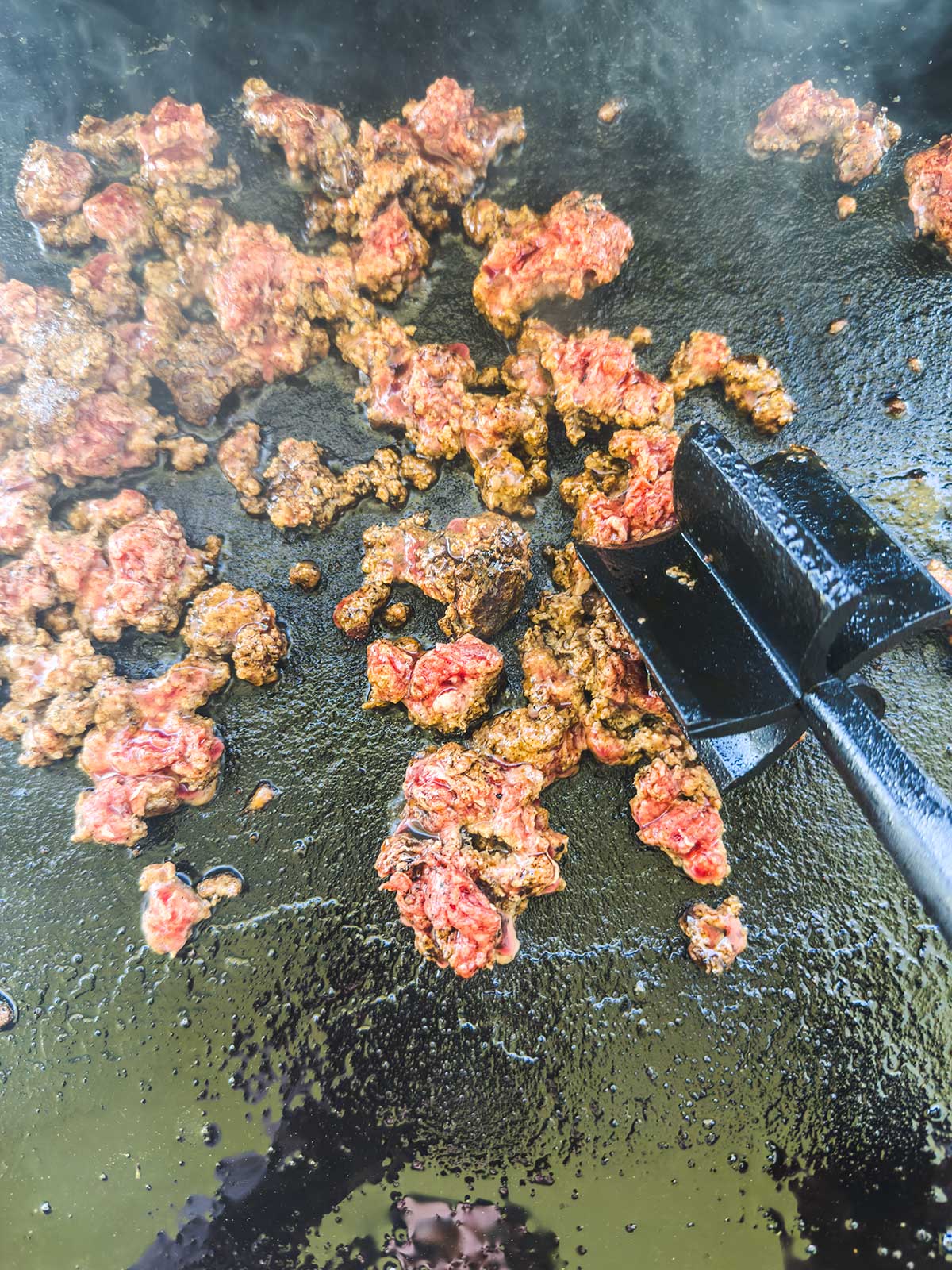 Ground beef is being cooked on a flat grill, like for a Blackstone egg roll in a bowl, with some pieces still red and raw, while others are browned. A metal spatula is pressing and breaking up the meat as it cooks.