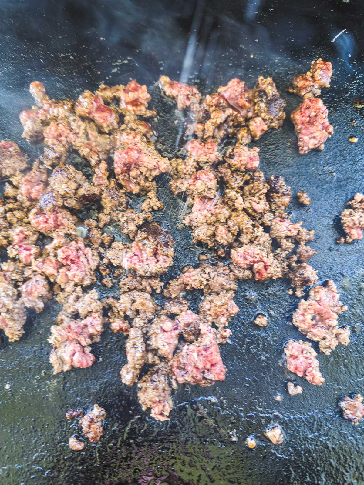 Ground beef is sizzling and browning on a Blackstone griddle, perfect for a blackstone egg roll in a bowl, with steam rising as some pieces remain pink while others are cooked through, creating an appetizing mix of textures and colors.