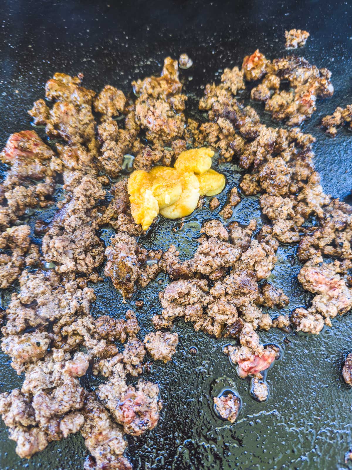 Ground beef cooking on a black griddle with yellow mustard and diced onions in the center, similar to making a Blackstone egg roll in a bowl. The beef is browned and slightly greasy.