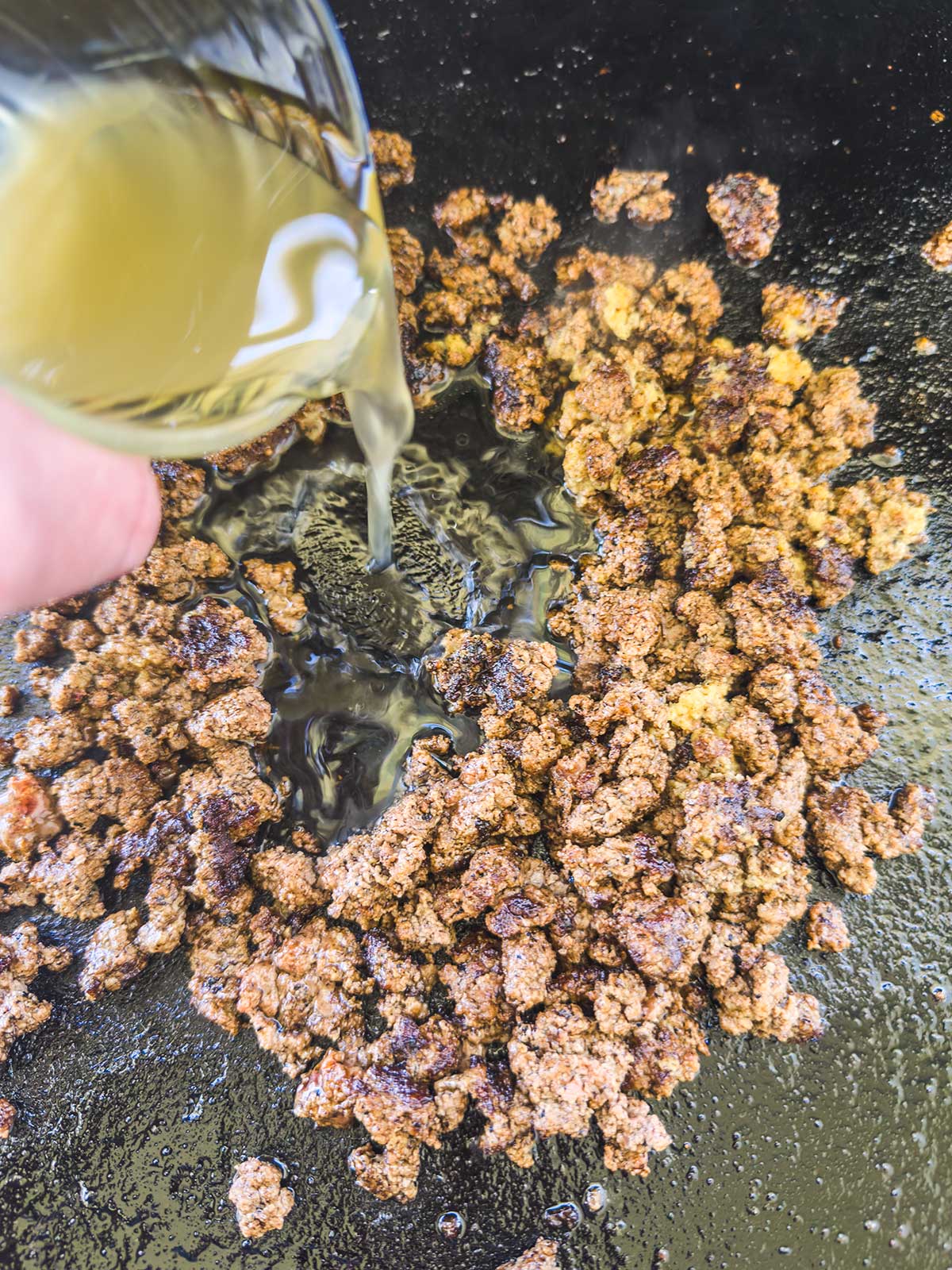 A hand pours liquid from a small glass container onto cooked ground beef in a skillet, a perfect step when making a Blackstone egg roll in a bowl.