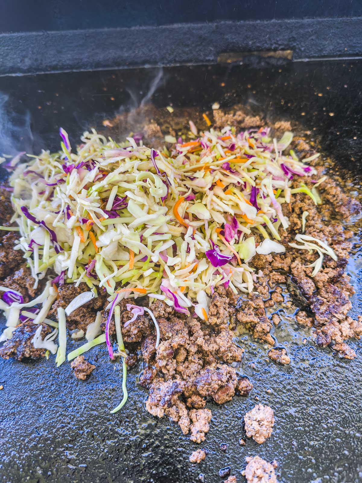 Ground beef and a colorful mix of shredded cabbage, carrots, and purple cabbage sizzle on the grill, creating a savory Blackstone egg roll in a bowl as steam rises from the hot surface.