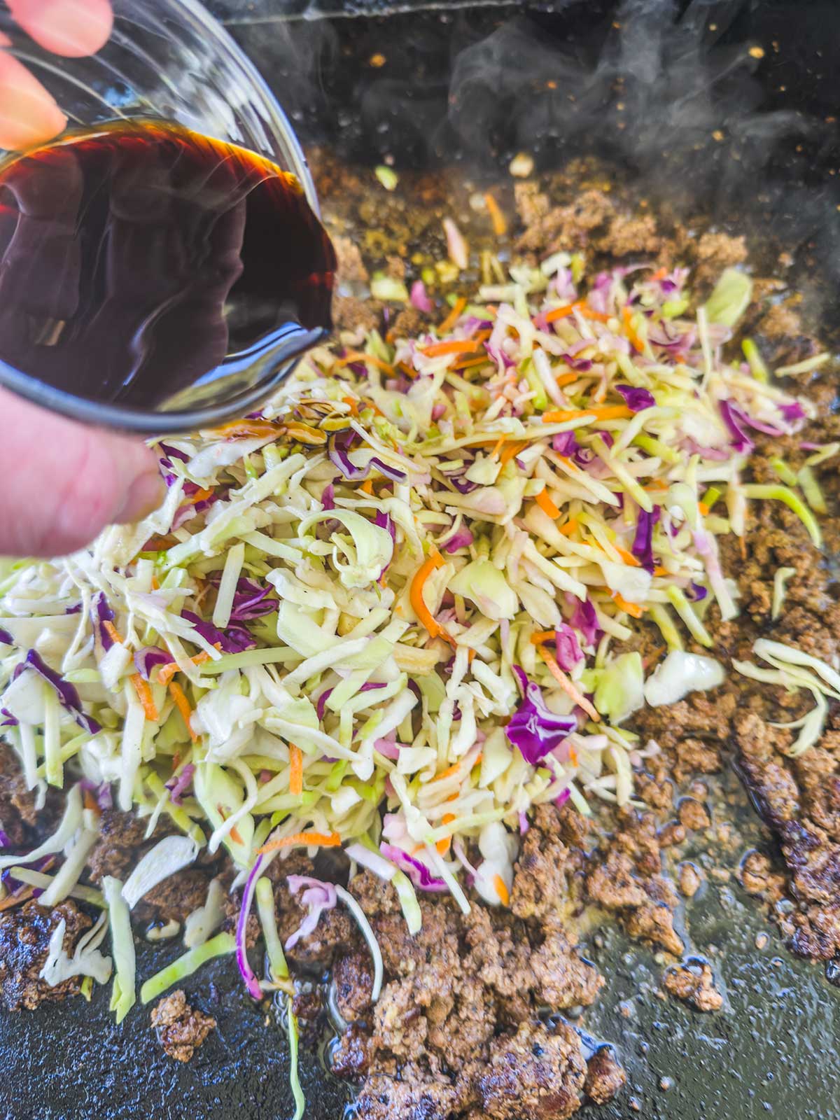 A hand pours dark soy sauce from a small glass bowl onto ground meat, shredded cabbage, carrots, and purple cabbage sizzling in a skillet—steam rises as this Blackstone egg roll in a bowl comes together.
