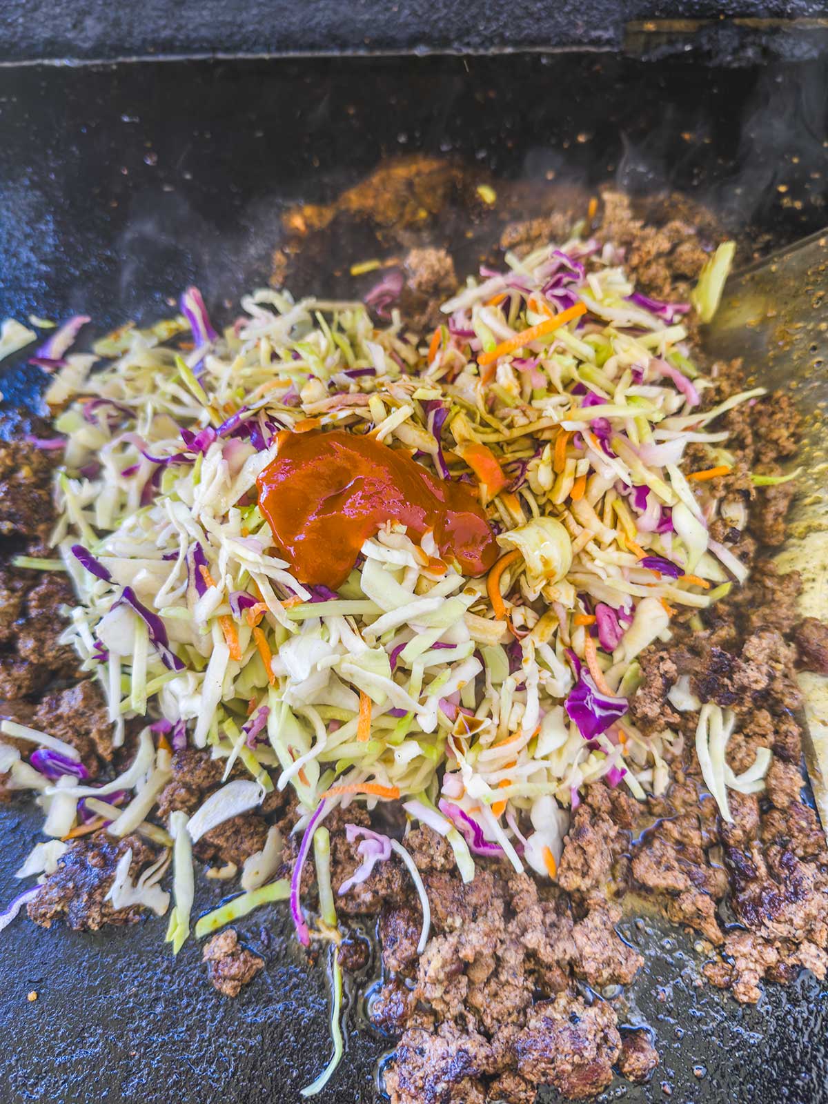 Ground beef sizzles on a griddle with shredded cabbage, carrots, and red cabbage for a delicious Blackstone egg roll in a bowl, topped with a dollop of red sauce and ready to be mixed together.