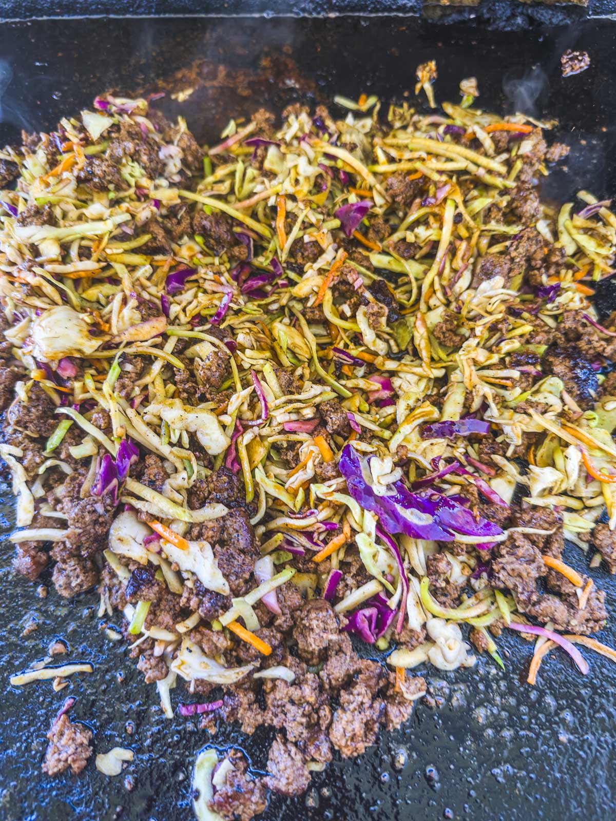 Ground beef and mixed shredded vegetables, including cabbage and carrots, sizzle on the griddle for a delicious Blackstone egg roll in a bowl. The ingredients are partially browned and full of flavor.