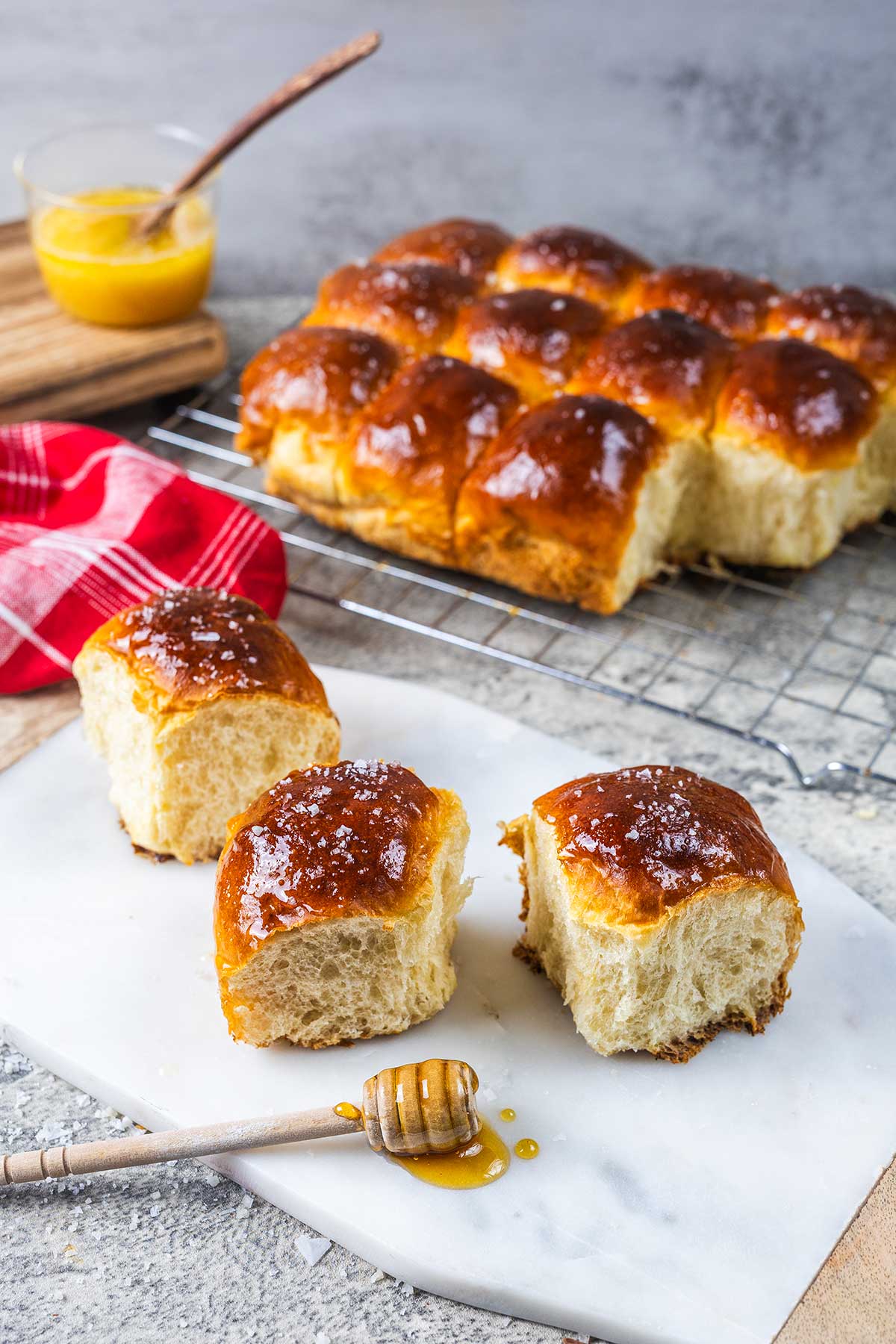 Three golden, fluffy yeast rolls with honey drizzle on a marble board, with more rolls in the background on a cooling rack. A honey dipper and a small jar of honey sit nearby.