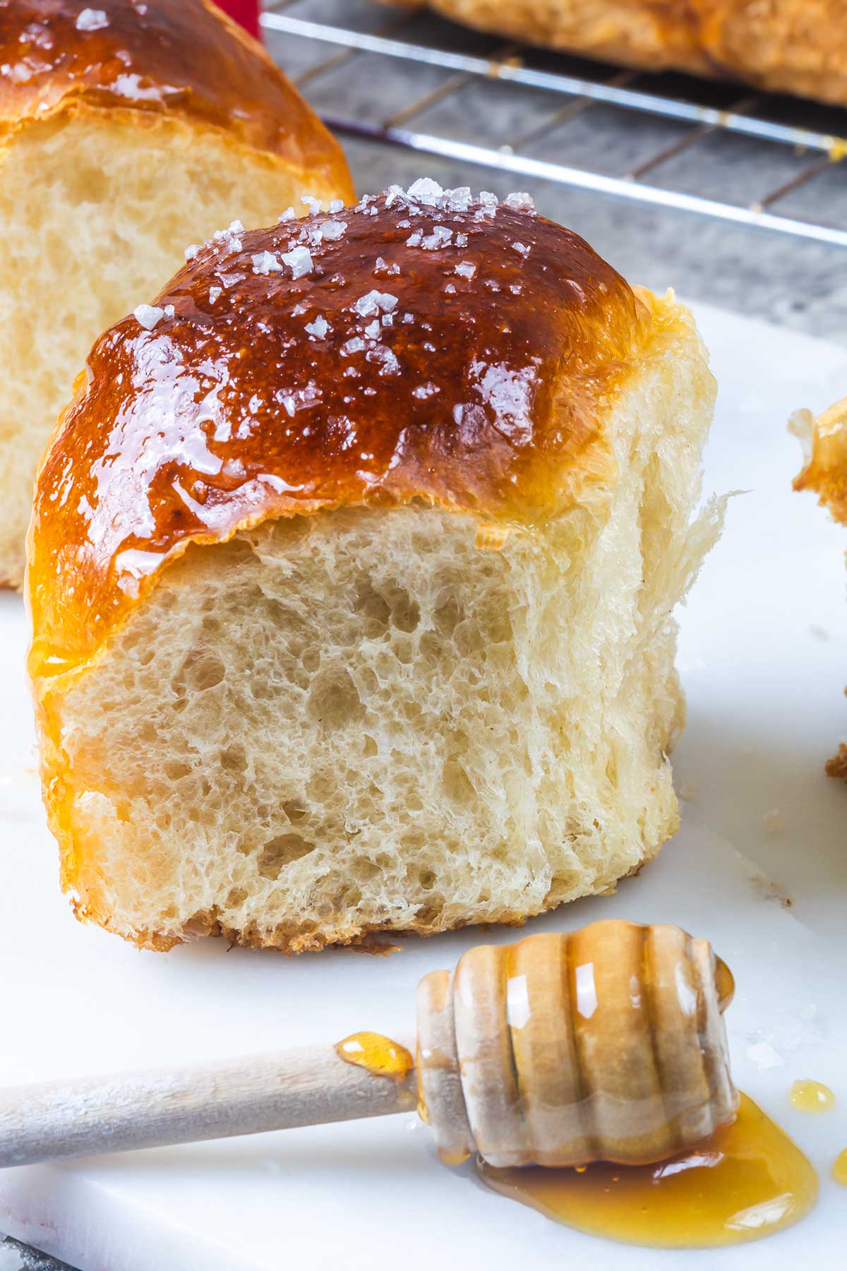 A close-up of a fluffy, golden-brown brioche roll sprinkled with coarse salt, next to a honey dipper with honey on a white surface. Another brioche is visible in the background.