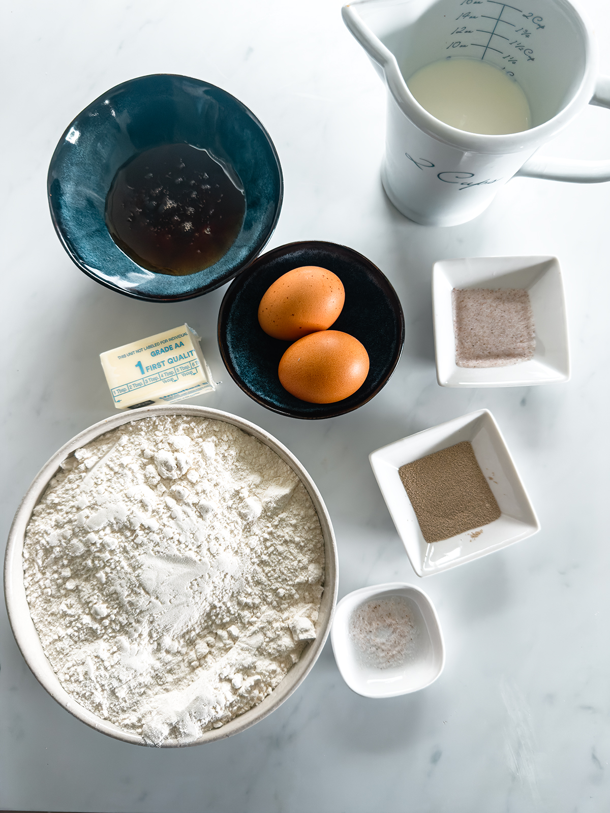 Overhead view of baking ingredients on a white surface: a bowl of flour, two eggs, a stick of butter, a blue bowl with a dark liquid, a small pitcher of milk, and small dishes containing yeast, salt, and sugar.