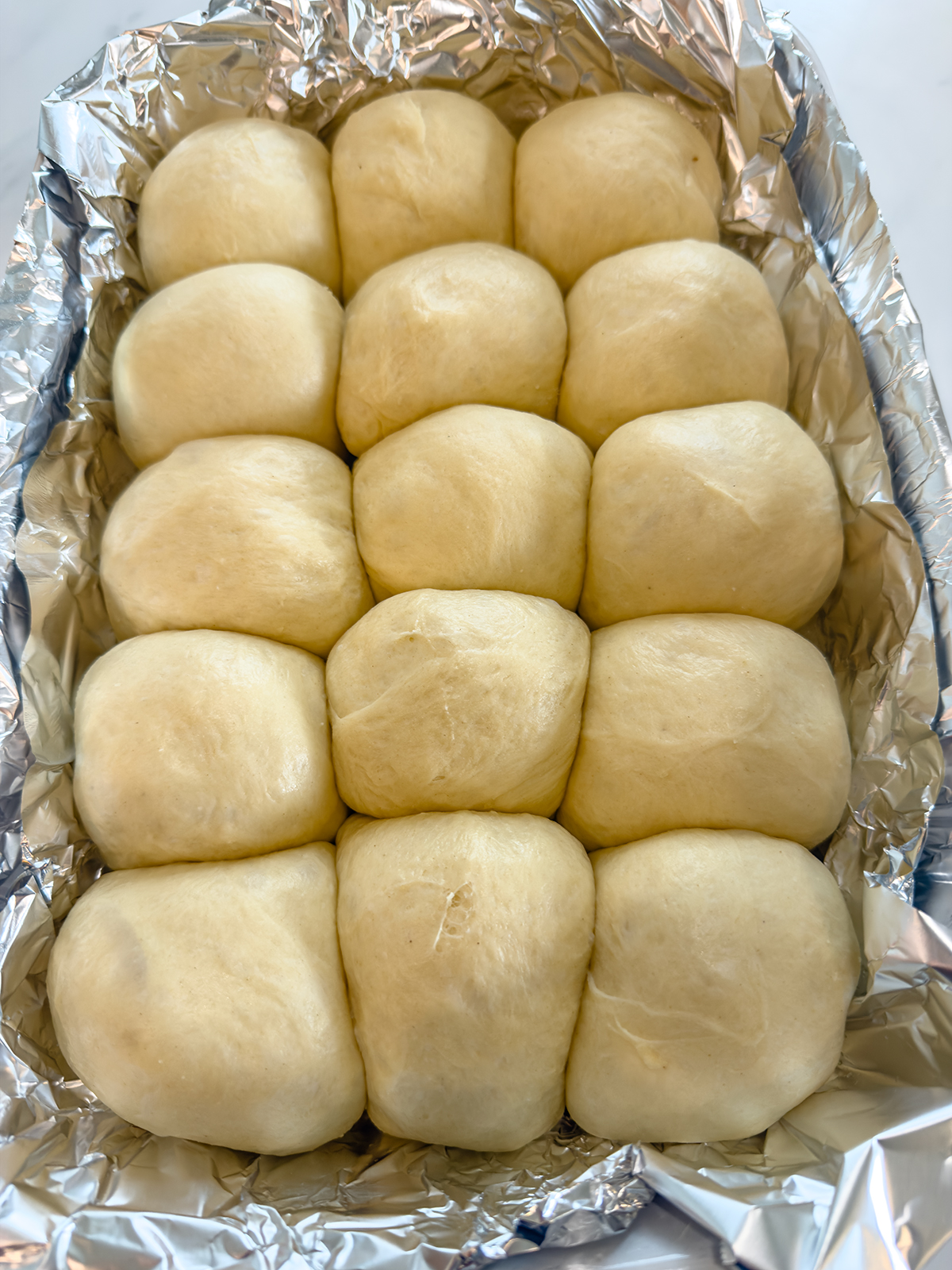 A baking dish lined with aluminum foil holds sixteen evenly arranged unbaked dough balls, ready to be baked into rolls.