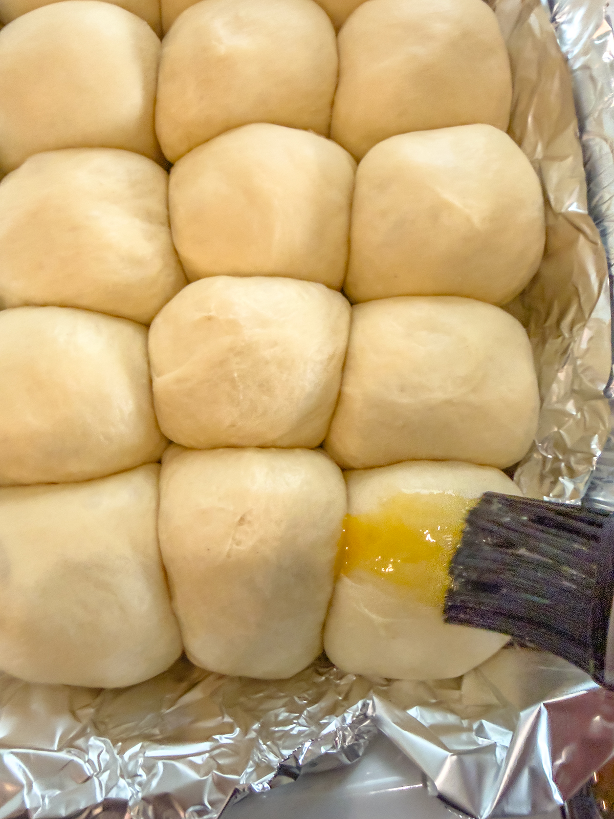A close-up of unbaked bread rolls in a foil-lined pan, with a pastry brush applying melted butter or egg wash to one of the rolls.