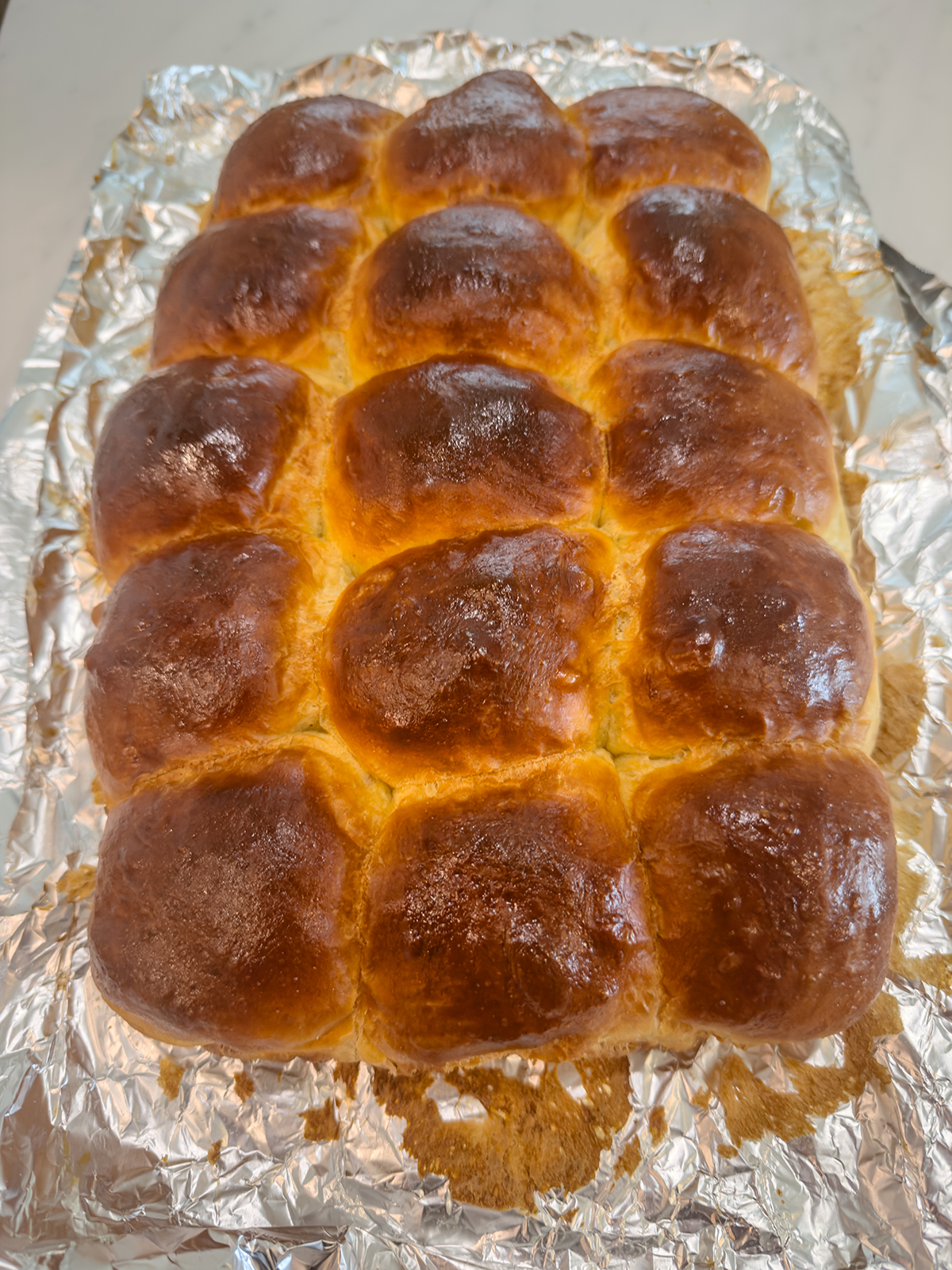 A batch of golden brown dinner rolls freshly baked, still closely packed together on a sheet of crumpled aluminum foil. The rolls have a shiny, glazed surface and are resting on a light-colored countertop.