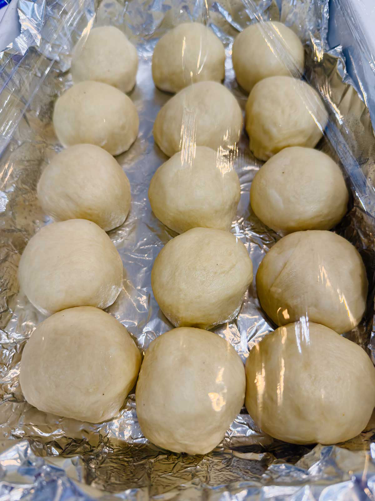 Fifteen round balls of dough are arranged in rows on a sheet of aluminum foil, covered loosely with plastic wrap, likely to rise before baking.