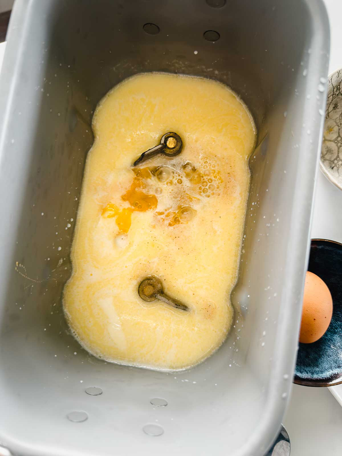 Eggs and beaten liquid, likely milk or egg mixture, are inside the metal bowl of a bread machine. A whole egg sits in a small dark bowl to the side. Bread machine kneading paddles are visible at the bottom.
