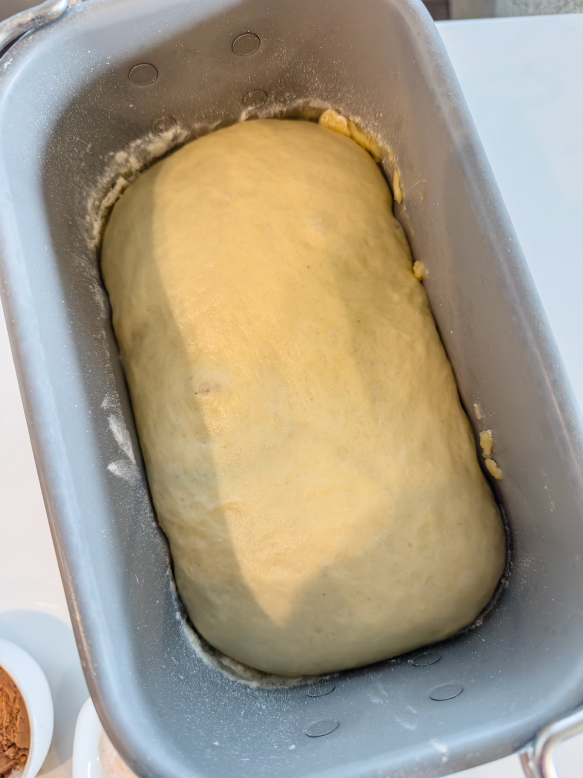 A loaf of risen bread dough in a rectangular metal bread machine pan, ready to be baked.