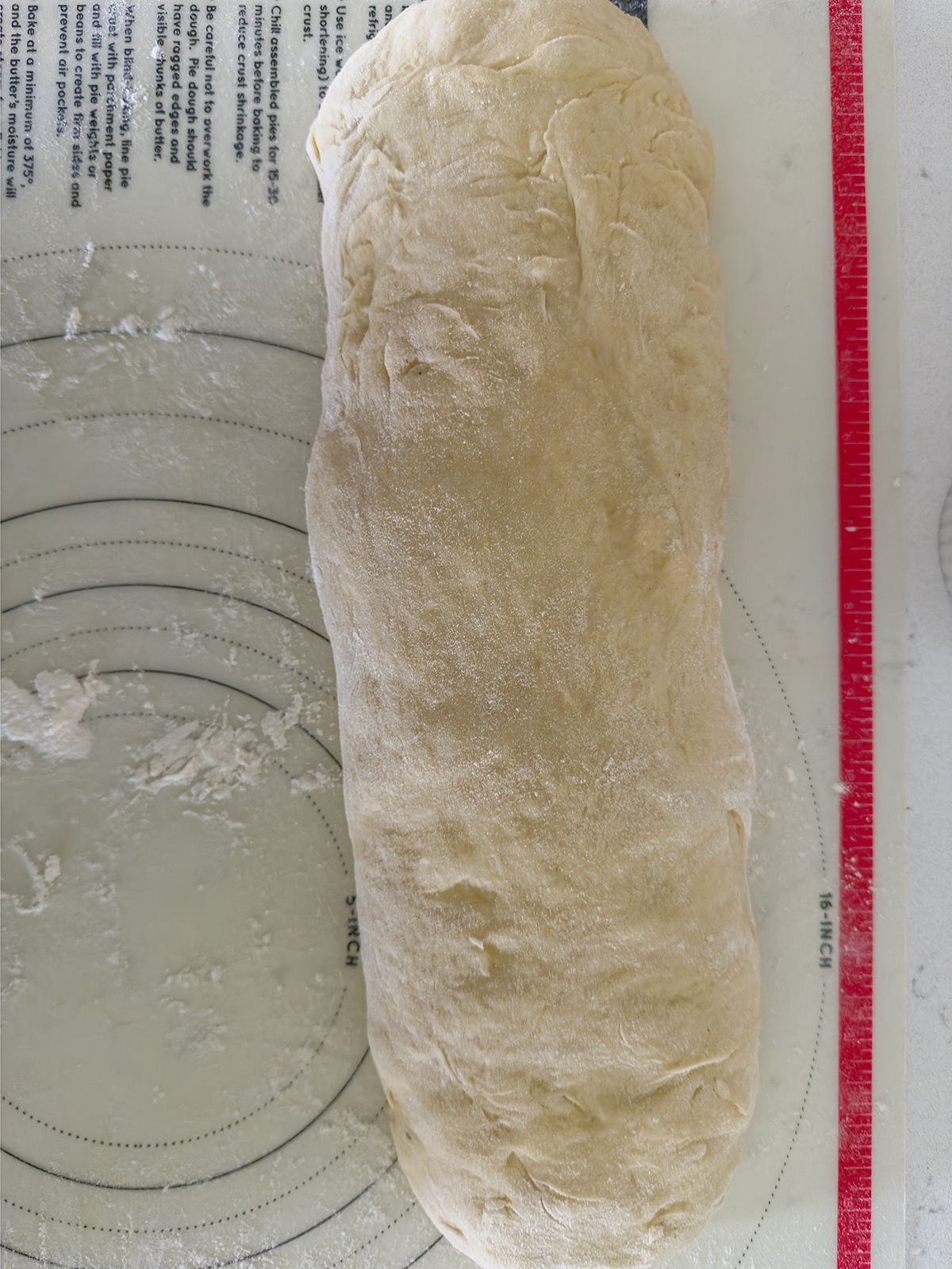 A lightly floured, folded loaf of bread dough rests on a silicone pastry mat with measurement markings and faint traces of flour.