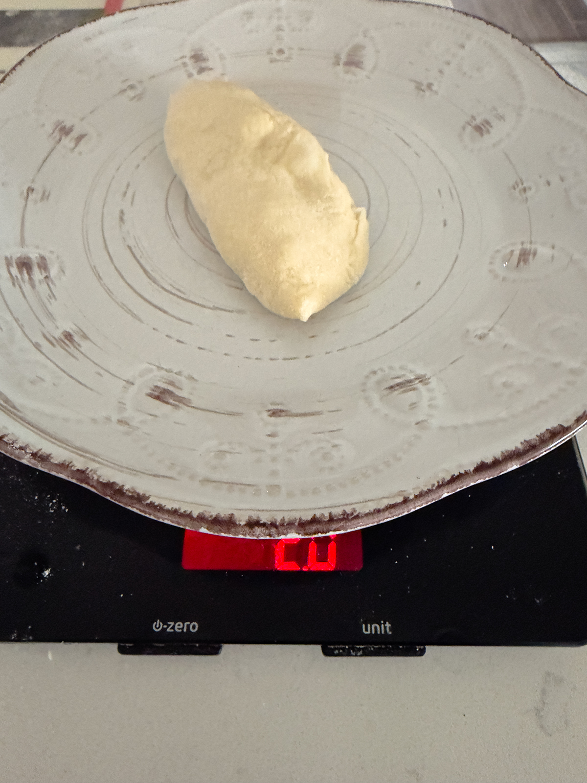 A piece of dough sits on a patterned plate, which is placed on a digital kitchen scale displaying a weight of 60 grams.