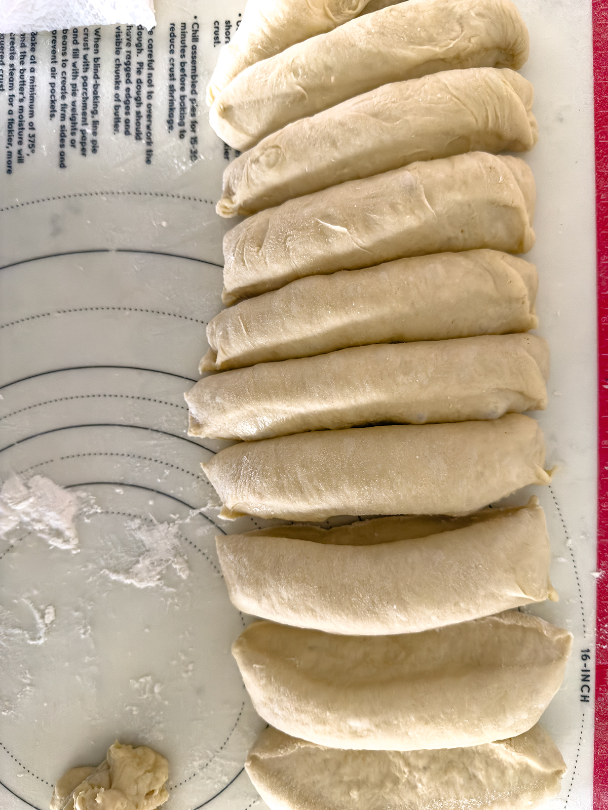 Nine evenly shaped pieces of dough are arranged in a row on a floured surface, ready for further preparation. A small scrap of leftover dough sits at the bottom left of the image.