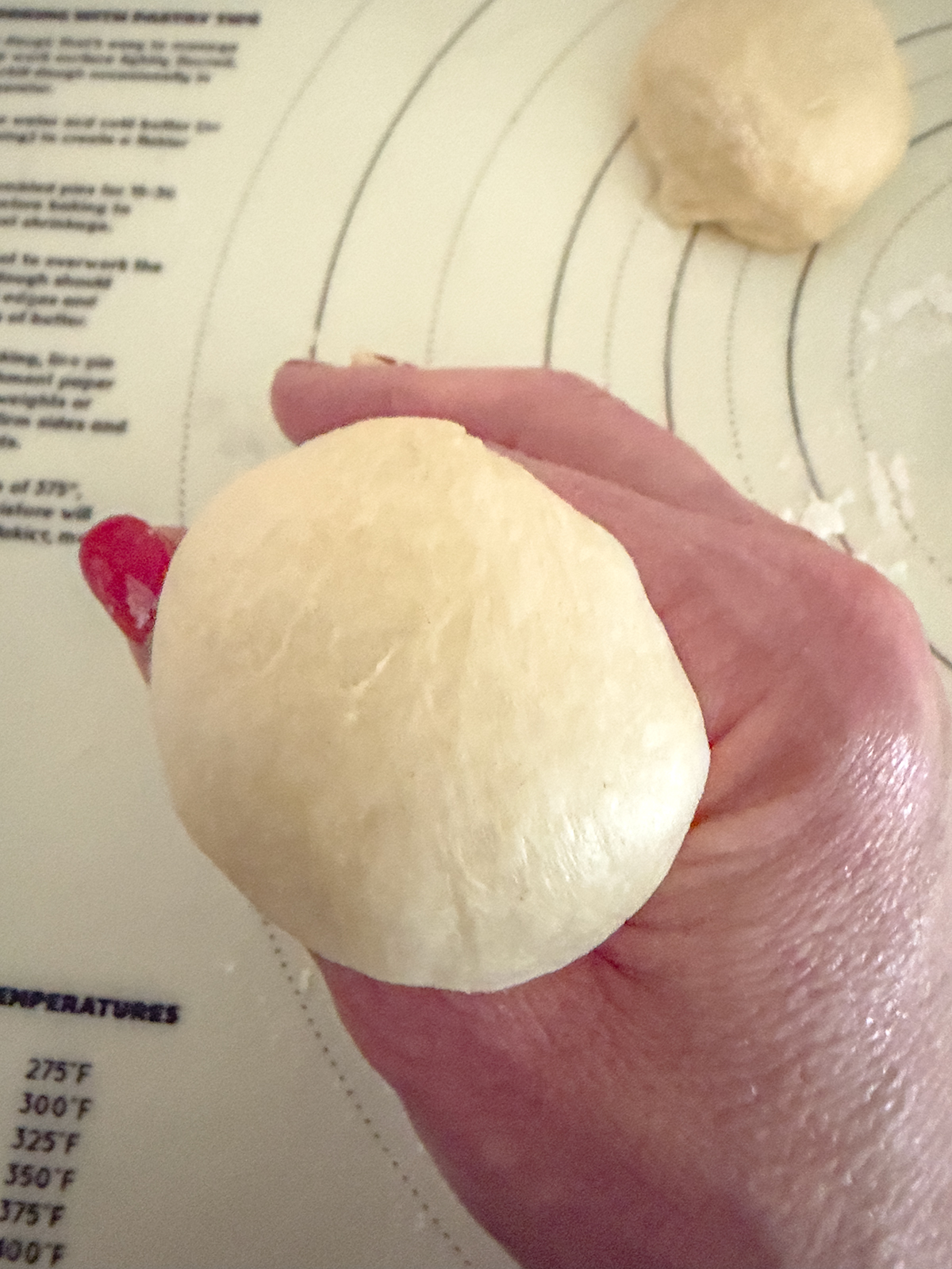 A hand holding a smooth ball of dough over a baking mat with temperature markings, with another ball of dough in the background.