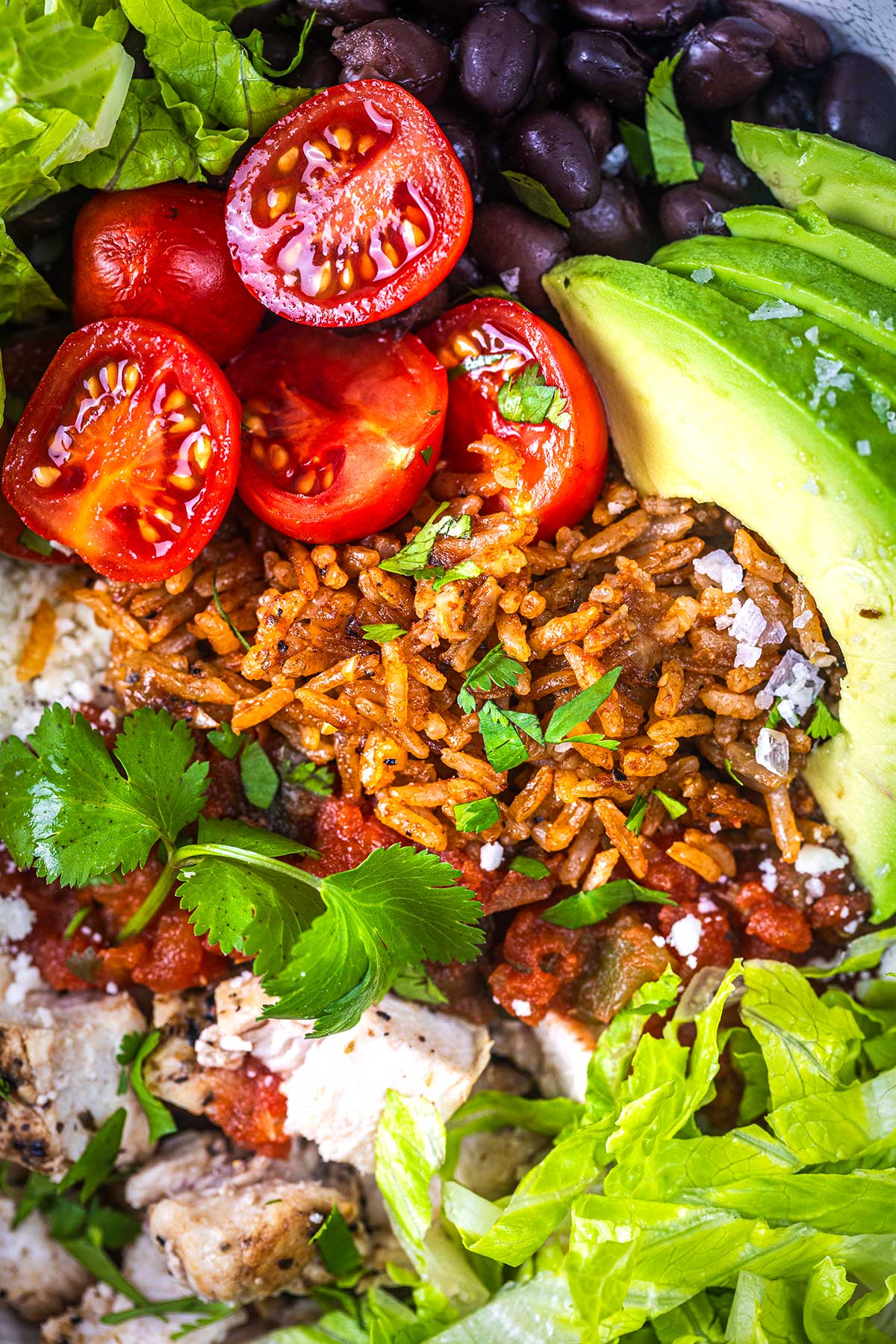 A close-up of a burrito bowl with black beans, sliced avocado, cherry tomatoes, seasoned rice, grilled chicken, lettuce, salsa, cilantro, and crumbled cheese.