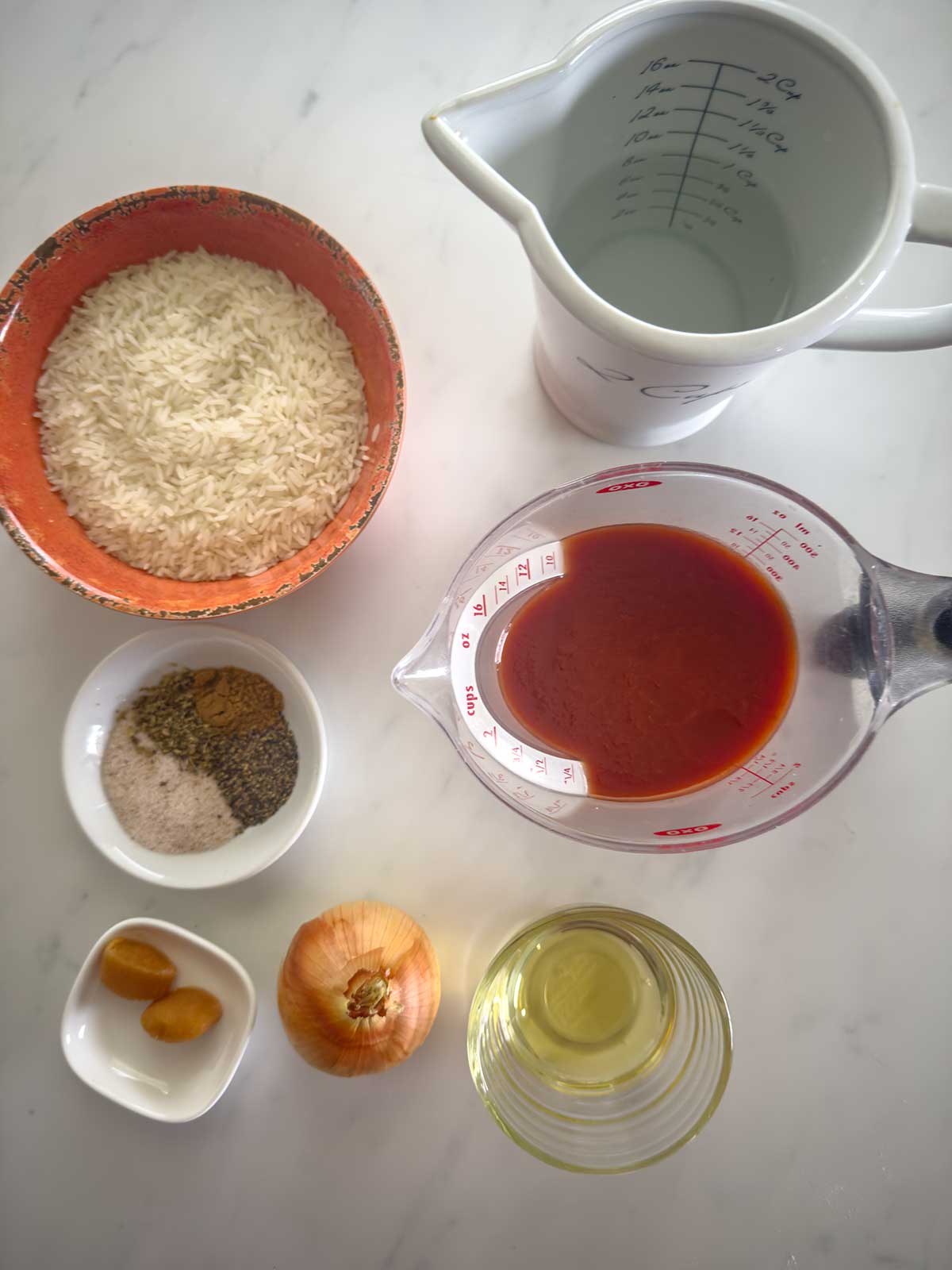 Top-down view of ingredients on a white surface: a bowl of rice, a measuring jug of water, a measuring cup of tomato sauce, small bowls of spices, garlic, an onion, and a small bowl of oil.