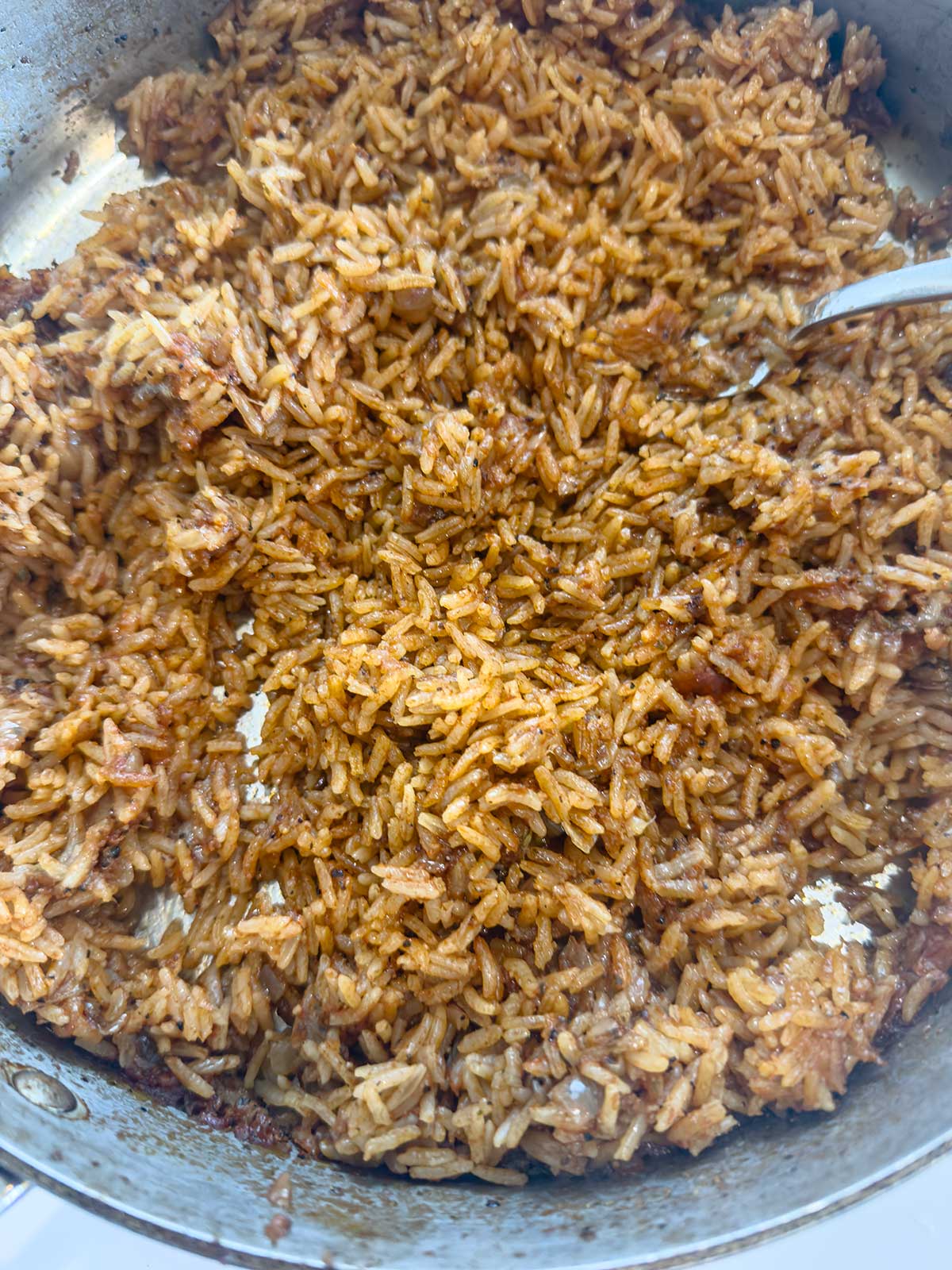 A close-up of a pan filled with cooked reddish-brown rice, likely seasoned or spiced, with a metal spoon partially visible on the right side.