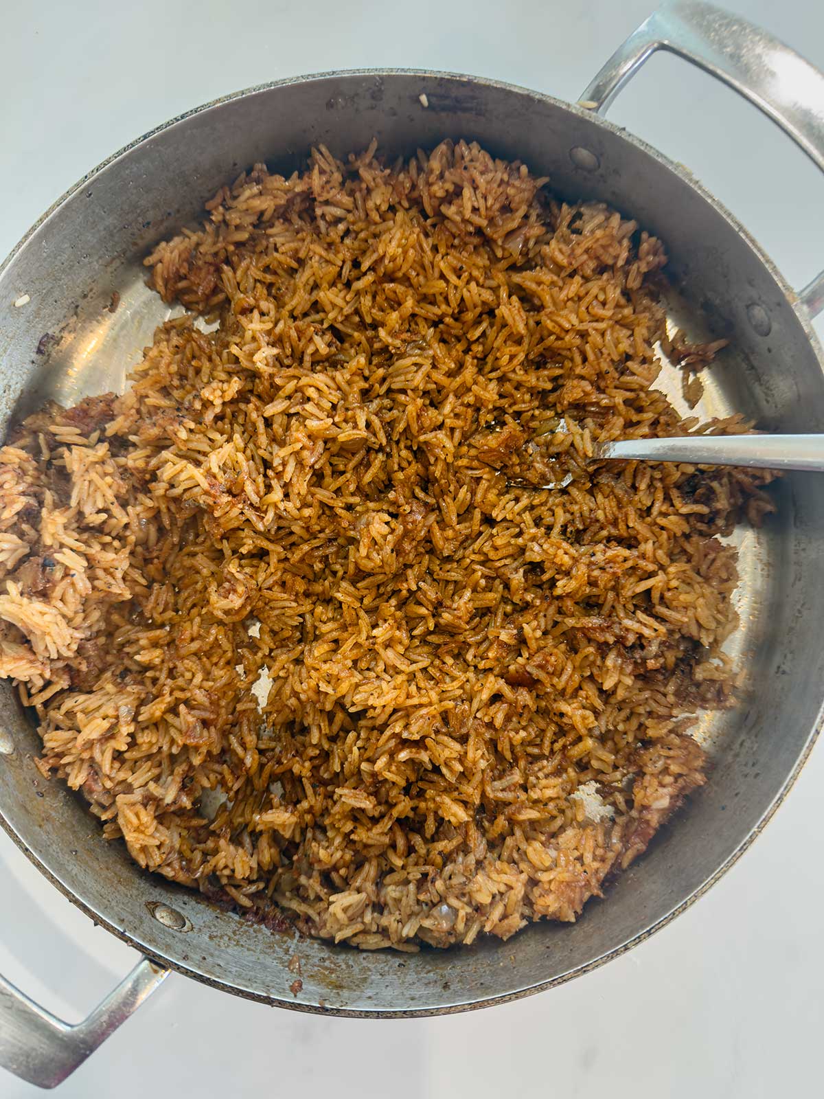 A silver pot filled with cooked, brown-colored rice, with a metal spoon partially submerged on the right side. The rice appears to be seasoned and slightly crispy in some areas.
