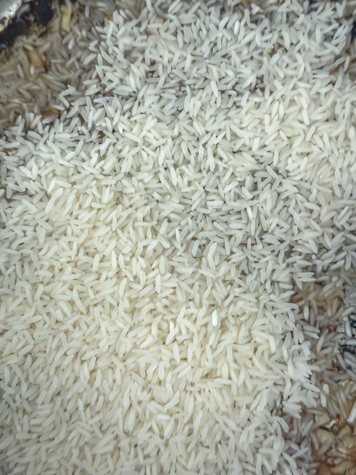 A close-up view of uncooked white rice grains scattered over a dark surface, with some brown and beige background elements partially visible.