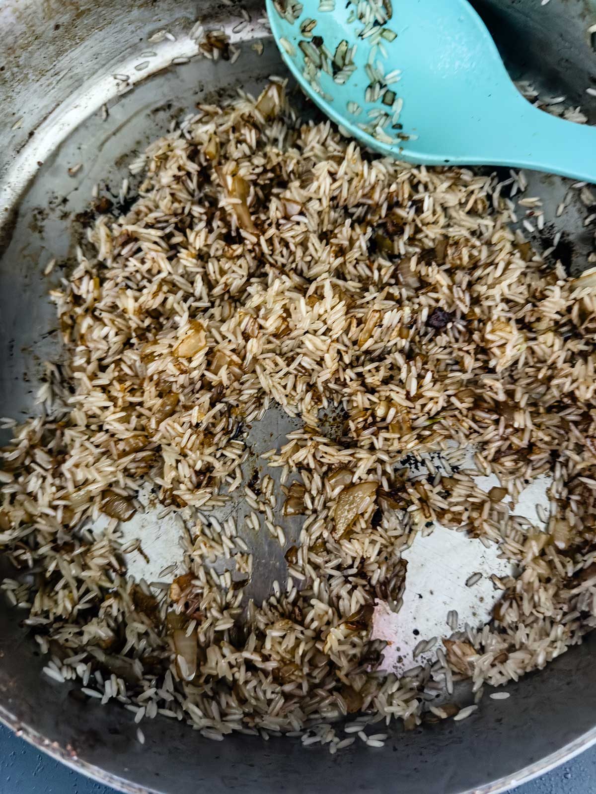 Uncooked rice and diced onions sautéed in a stainless steel pan, with some grains browned, and a blue spoon resting on the side.