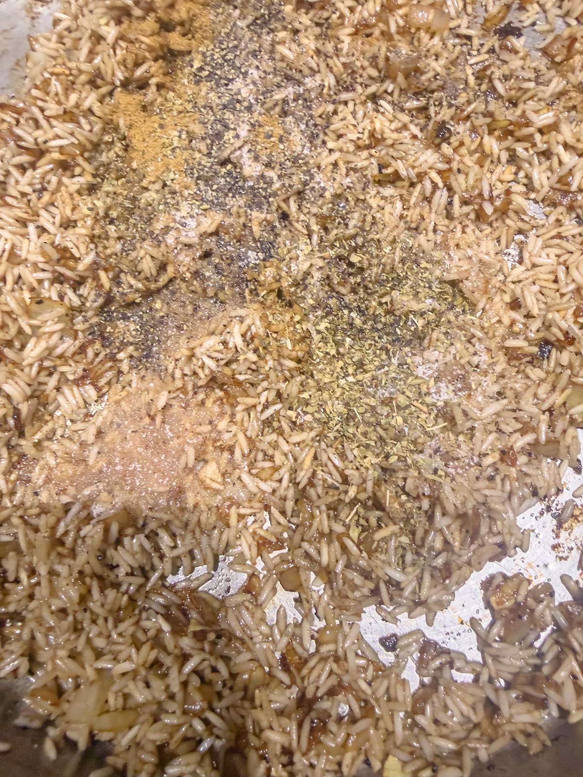 Uncooked rice mixed with browned onions, various dried herbs, and ground spices in a pan, ready to be cooked.