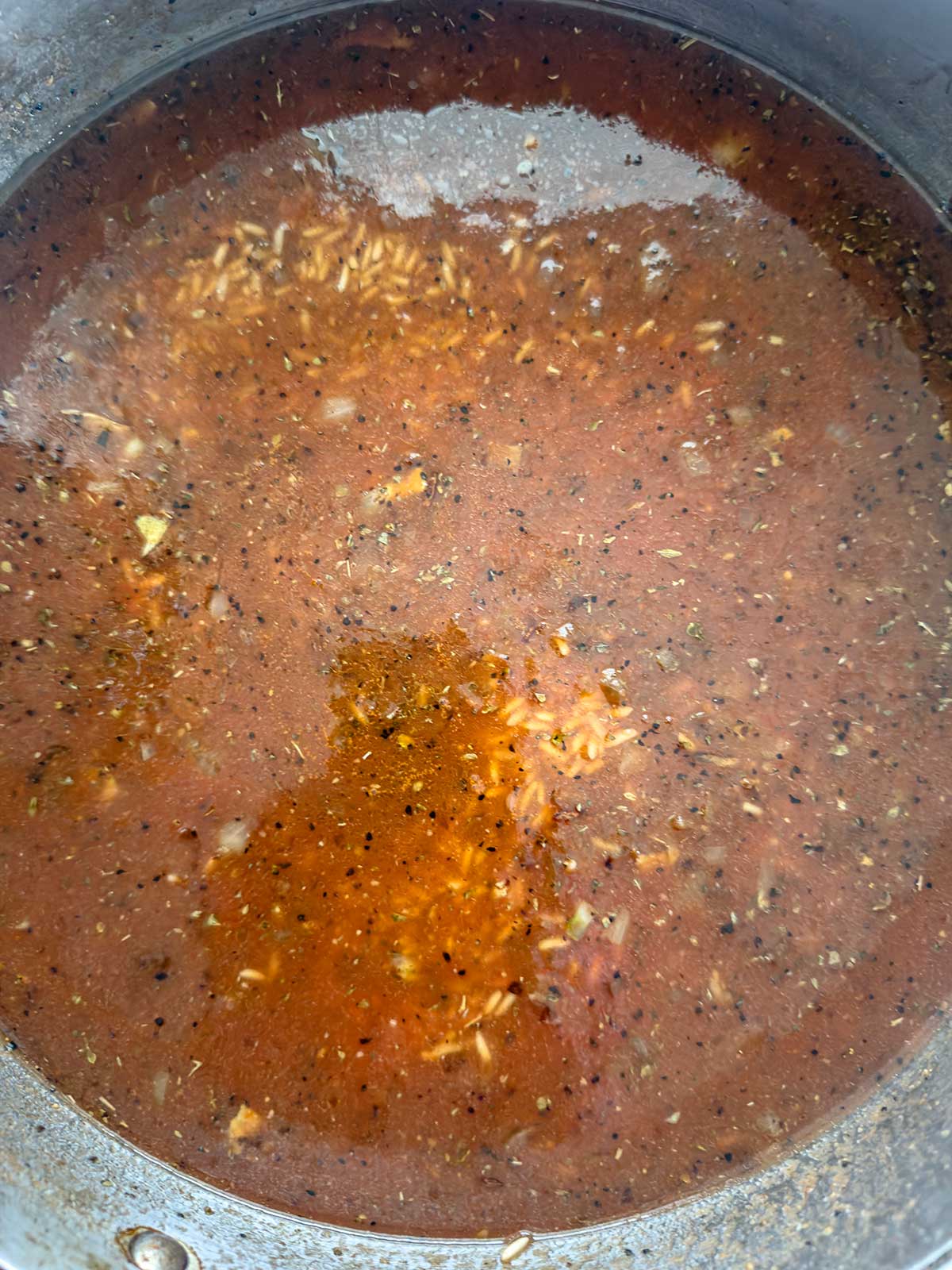 A pot filled with reddish-brown broth, floating herbs, spices, rice grains, and chopped vegetables, creating a hearty and flavorful soup.
