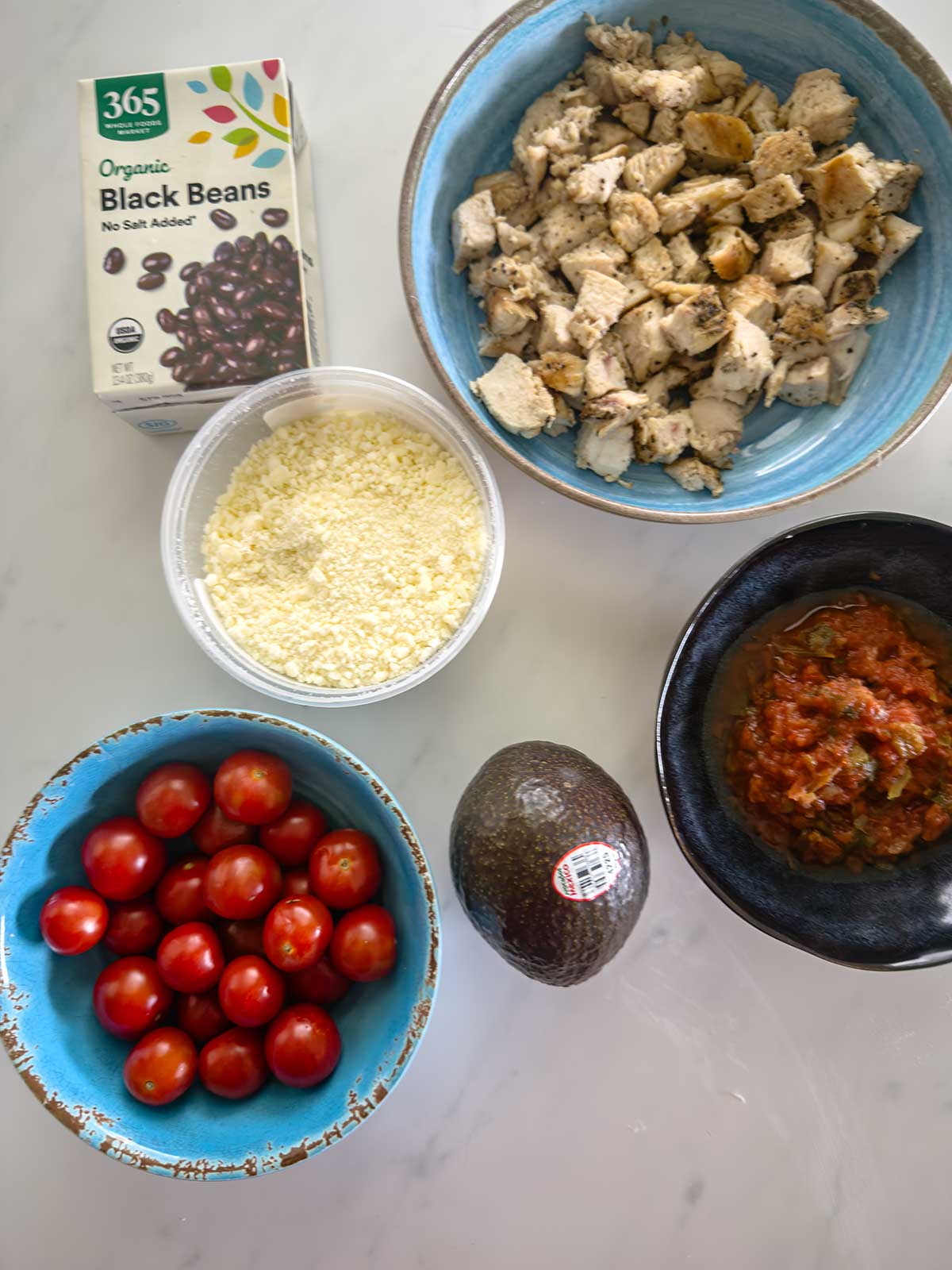 A marble surface with a carton of black beans, a bowl of cooked diced chicken, a bowl of shredded cheese, a bowl of salsa, a bowl of cherry tomatoes, and a whole avocado.