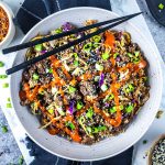 Egg roll in a bowl with ground meat, chopped green onions, shredded cabbage, sesame seeds, and a drizzle of red sauce, served with black chopsticks on top.