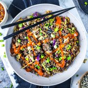 Egg roll in a bowl with ground meat, chopped green onions, shredded cabbage, sesame seeds, and a drizzle of red sauce, served with black chopsticks on top.