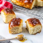 Three shiny, golden-brown brioche rolls sit on a marble board, sprinkled with coarse salt. A honey dipper with honey is nearby, and more brioche is cooling on a wire rack in the background.