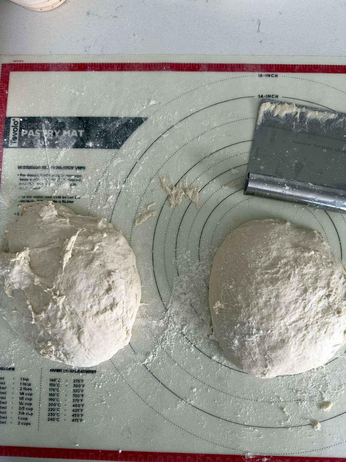 Two balls of bread dough on a red and white pastry mat with measurement markings; a metal bench scraper lies nearby, and flour is scattered on the surface.