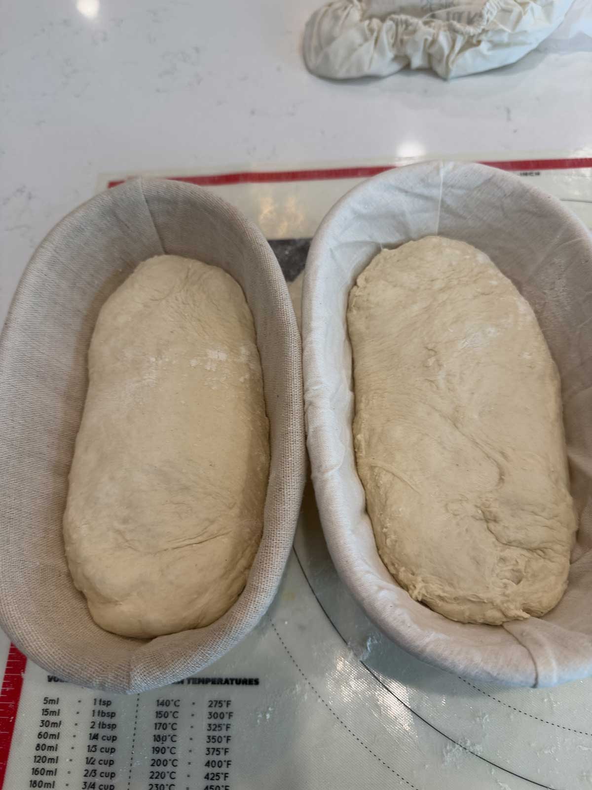Two oval-shaped dough loaves resting in cloth-lined proofing baskets on a kitchen counter, with a red and white baking mat and part of a cloth visible in the background.