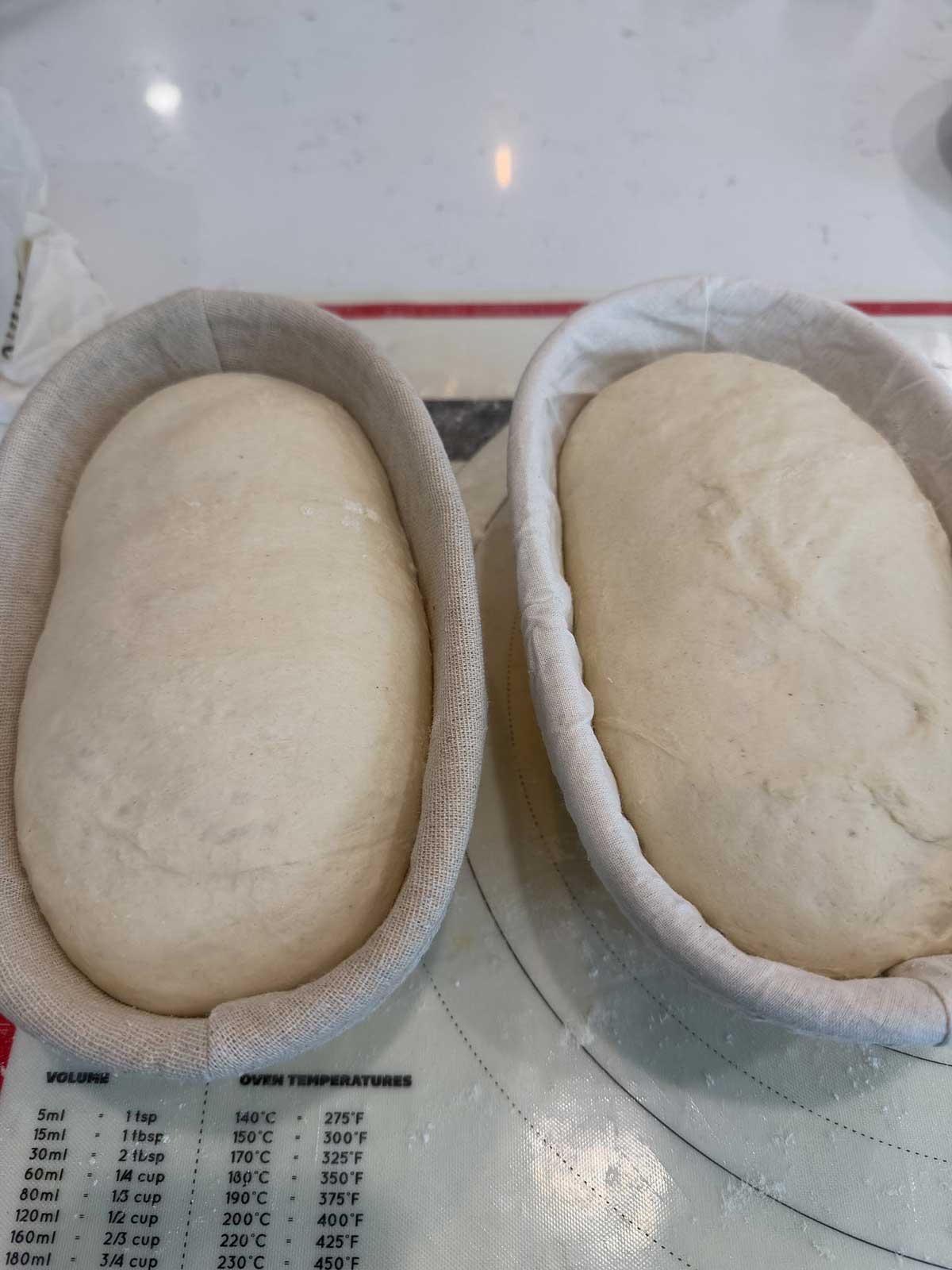 Two oval-shaped dough loaves rising in cloth-lined proofing baskets on a kitchen counter, with a baking temperature and measurement chart visible beneath them.