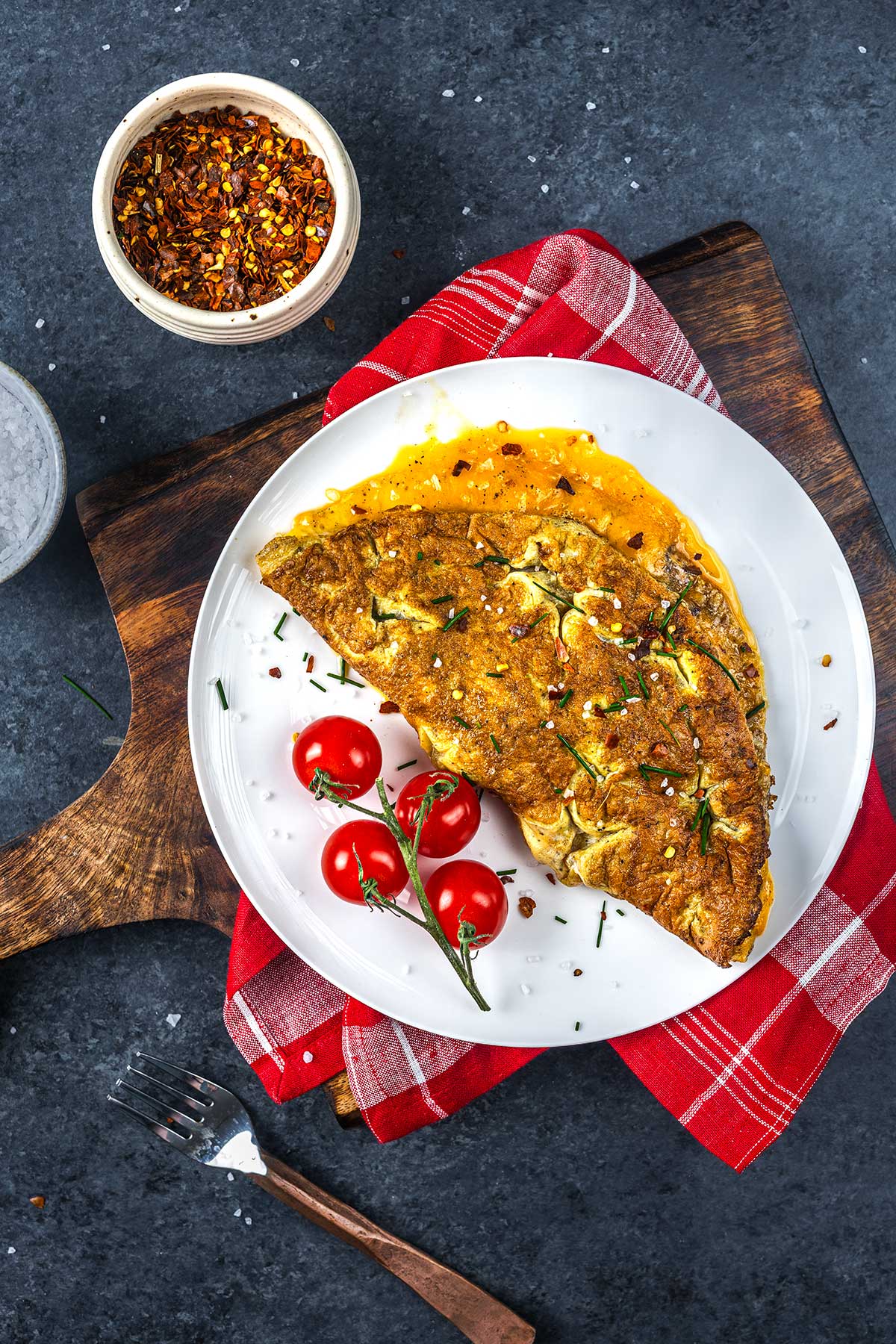 A golden-brown omelet garnished with herbs is served on a white plate alongside three vine tomatoes. The plate rests on a wooden board with a red plaid napkin. A small bowl of chili flakes is nearby.