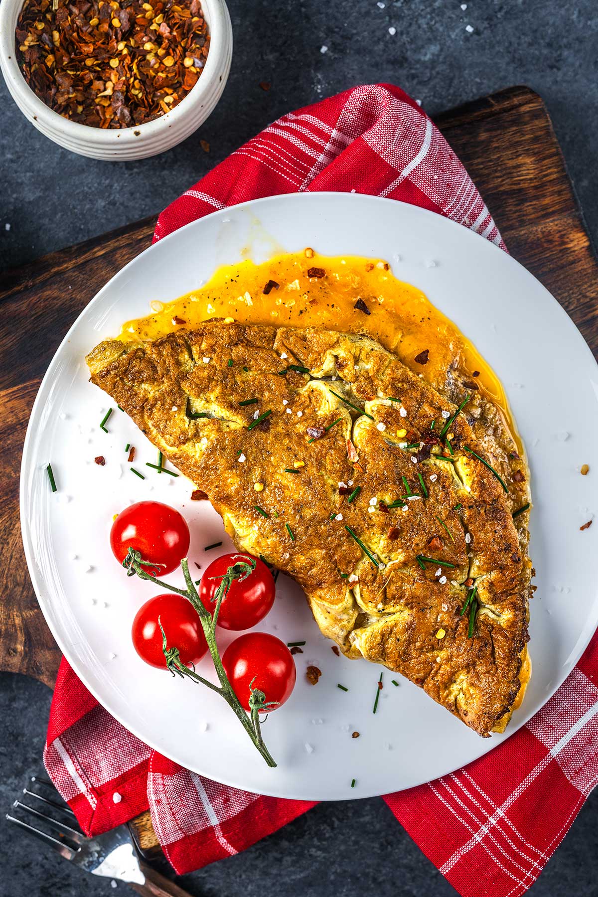 A golden-brown omelette garnished with herbs and chili flakes sits on a white plate next to vine tomatoes. The plate rests on a red plaid napkin atop a wooden board, with a small bowl of chili flakes nearby.