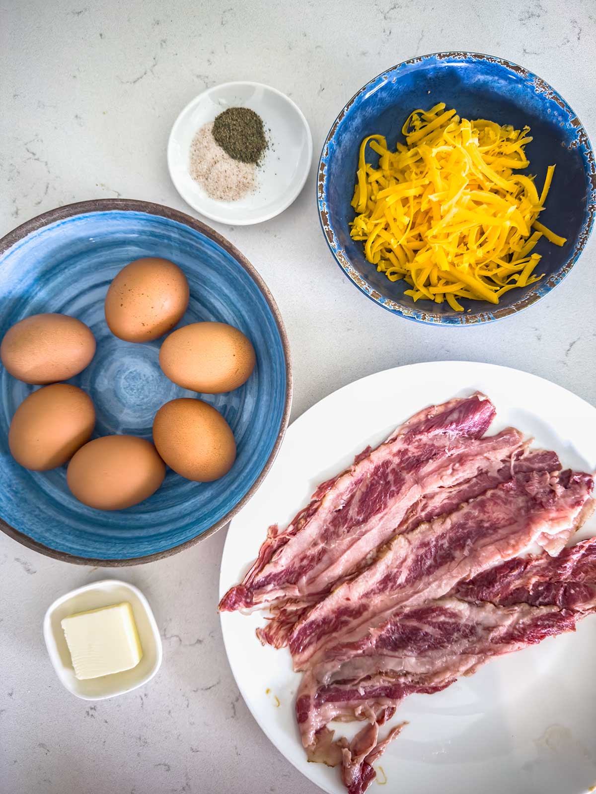 A top view of raw bacon strips on a plate, a bowl of shredded cheddar cheese, five eggs in a blue bowl, a dish of salt and pepper, and a small dish of butter on a light countertop.