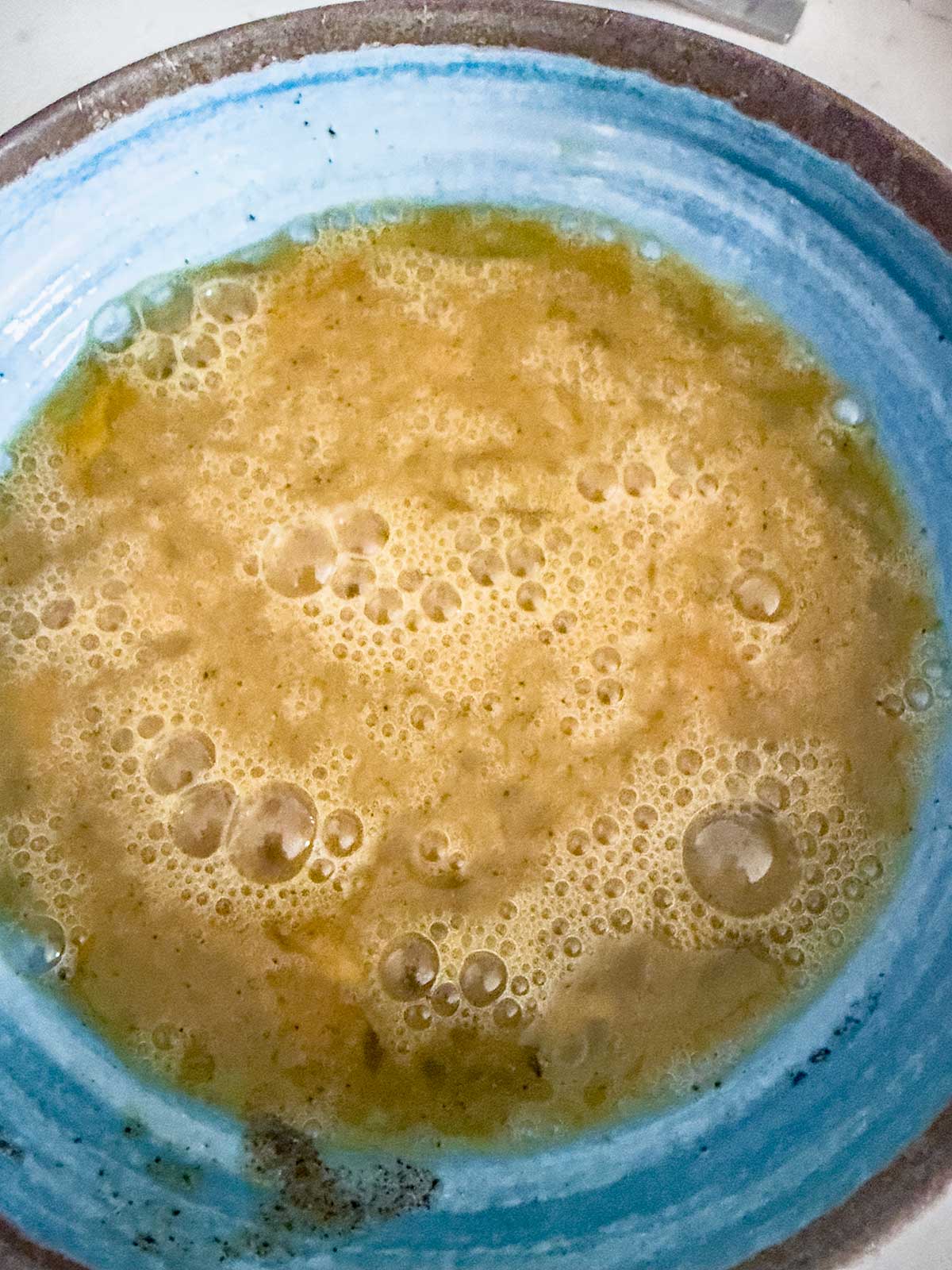 A close-up of a blue ceramic bowl filled with a yellow, frothy liquid that appears to be beaten eggs, with bubbles visible on the surface.